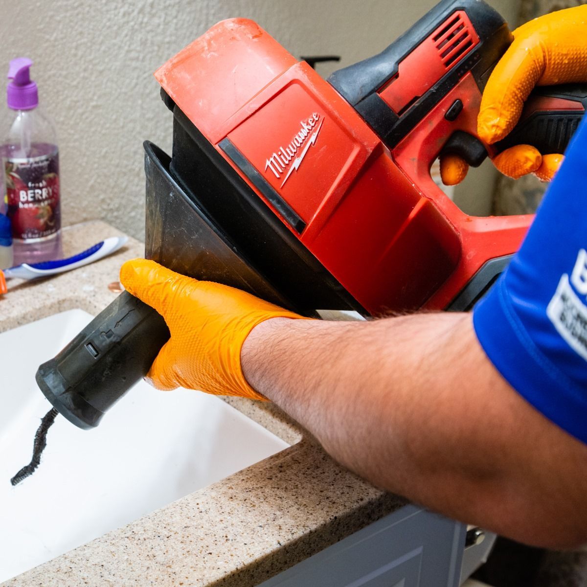 Person in orange gloves using a red Milwaukee drain cleaning machine to unclog a bathroom sink.