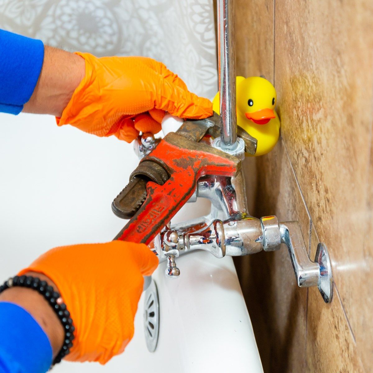 Plumber in orange gloves using a wrench on a faucet, yellow rubber duck, bathroom setting.