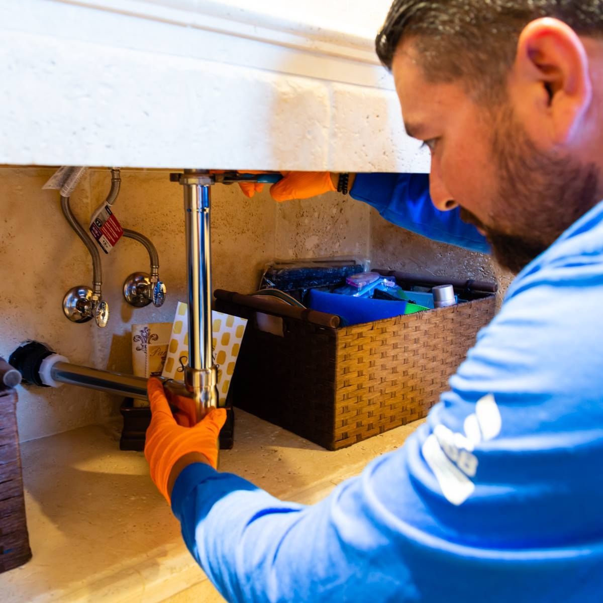 Plumber in blue shirt and orange gloves, working on pipes under a sink.
