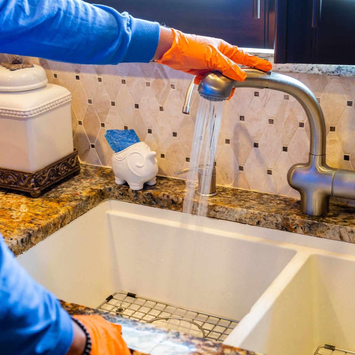 Person in orange gloves washing dishes in a white kitchen sink.