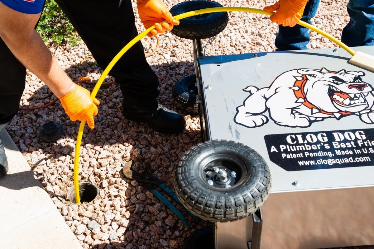 Two people in orange gloves feed a yellow drain snake into a hole, near a rolling 