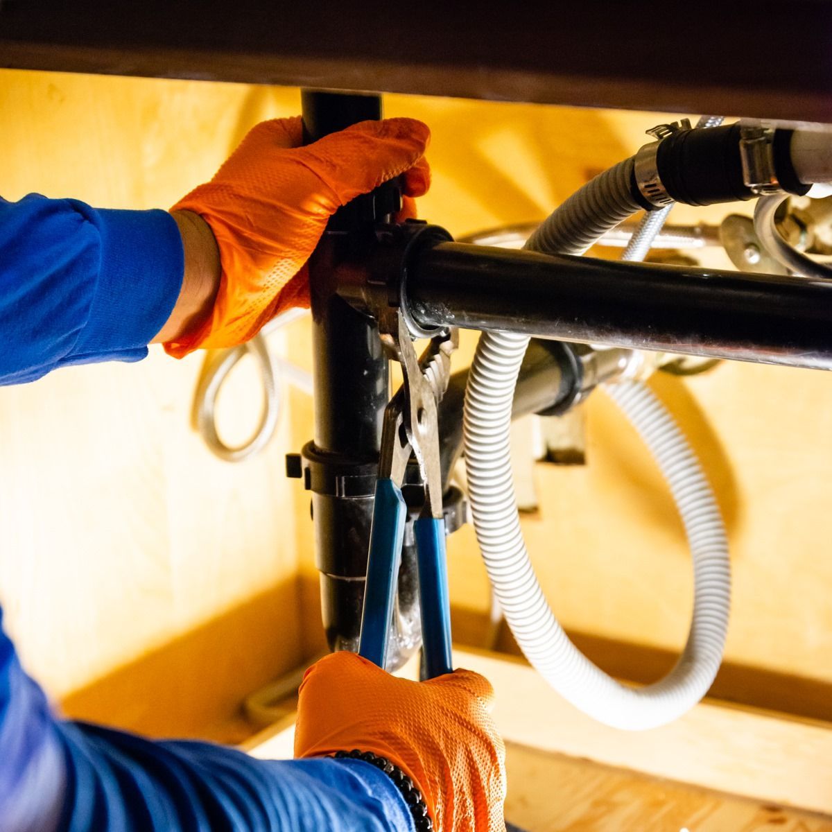 Person in orange gloves uses pliers to adjust pipes under a sink.