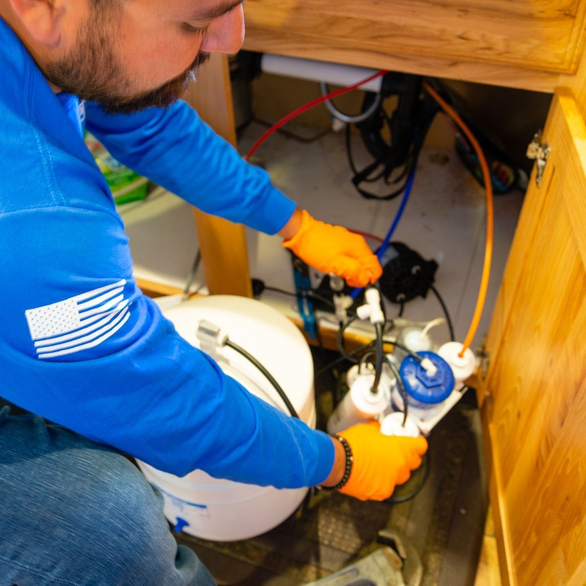 Man in orange gloves working on reverse osmosis water filtration system under a cabinet.