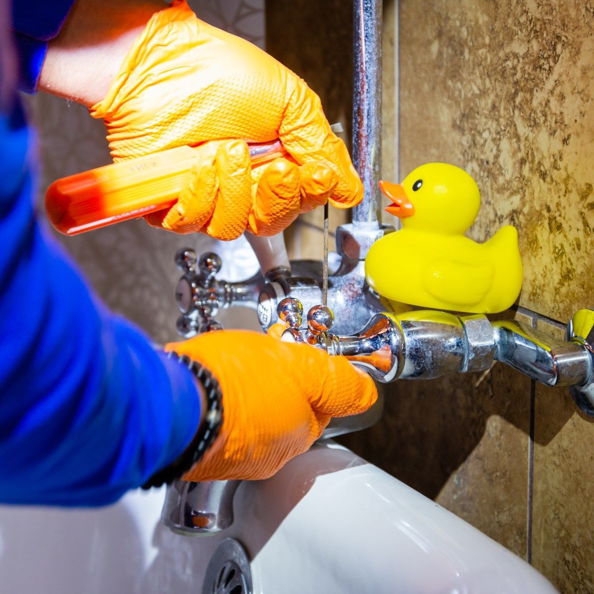 Person wearing orange gloves fixing bathroom faucet with a yellow rubber duck nearby.
