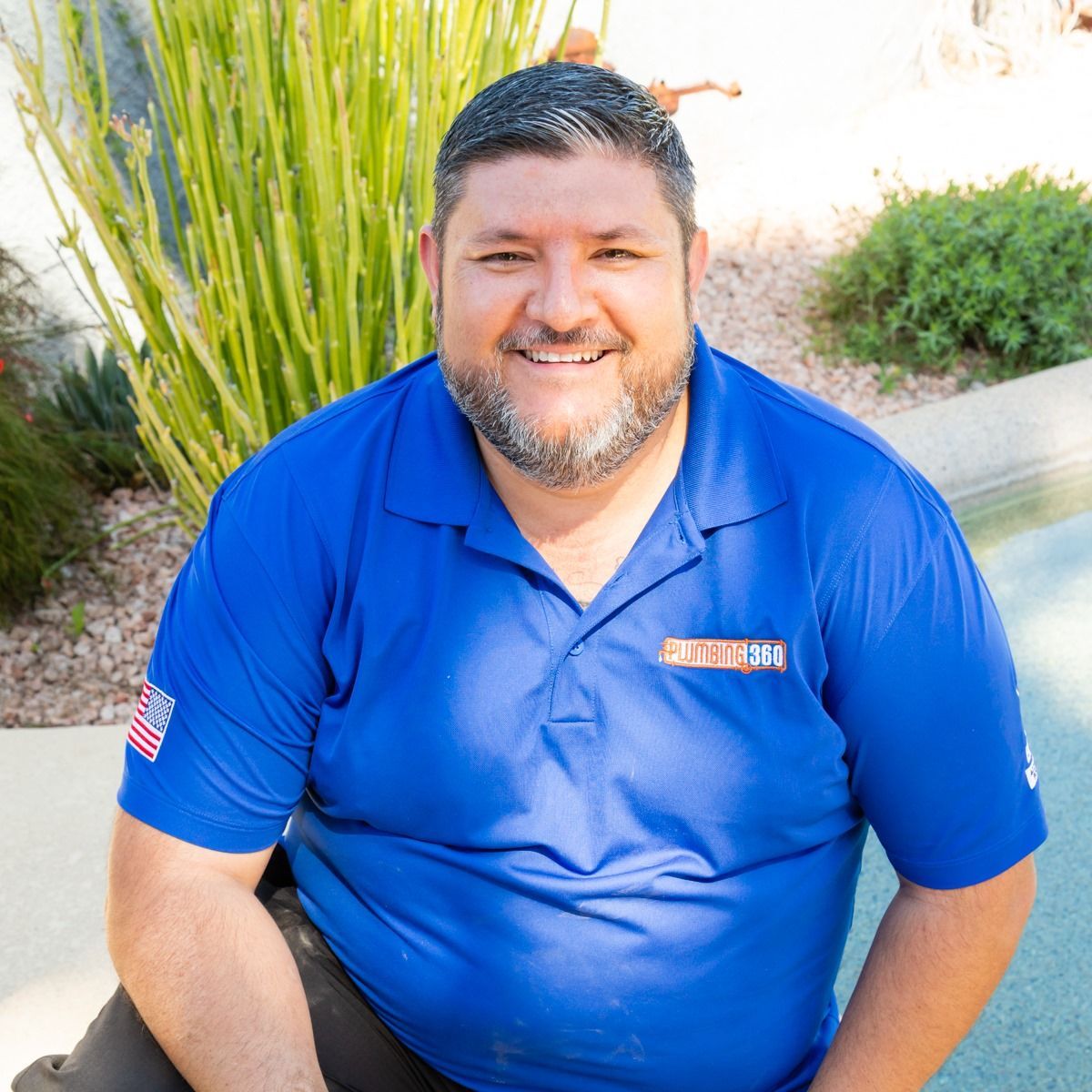 Man in blue polo shirt smiling, seated near green plants, sunny outdoor setting.