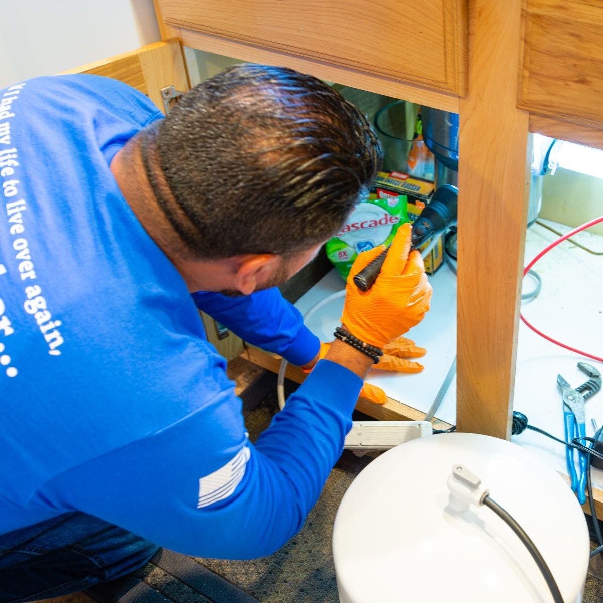 Plumber working under sink with orange gloves, holding flashlight, in a bathroom.