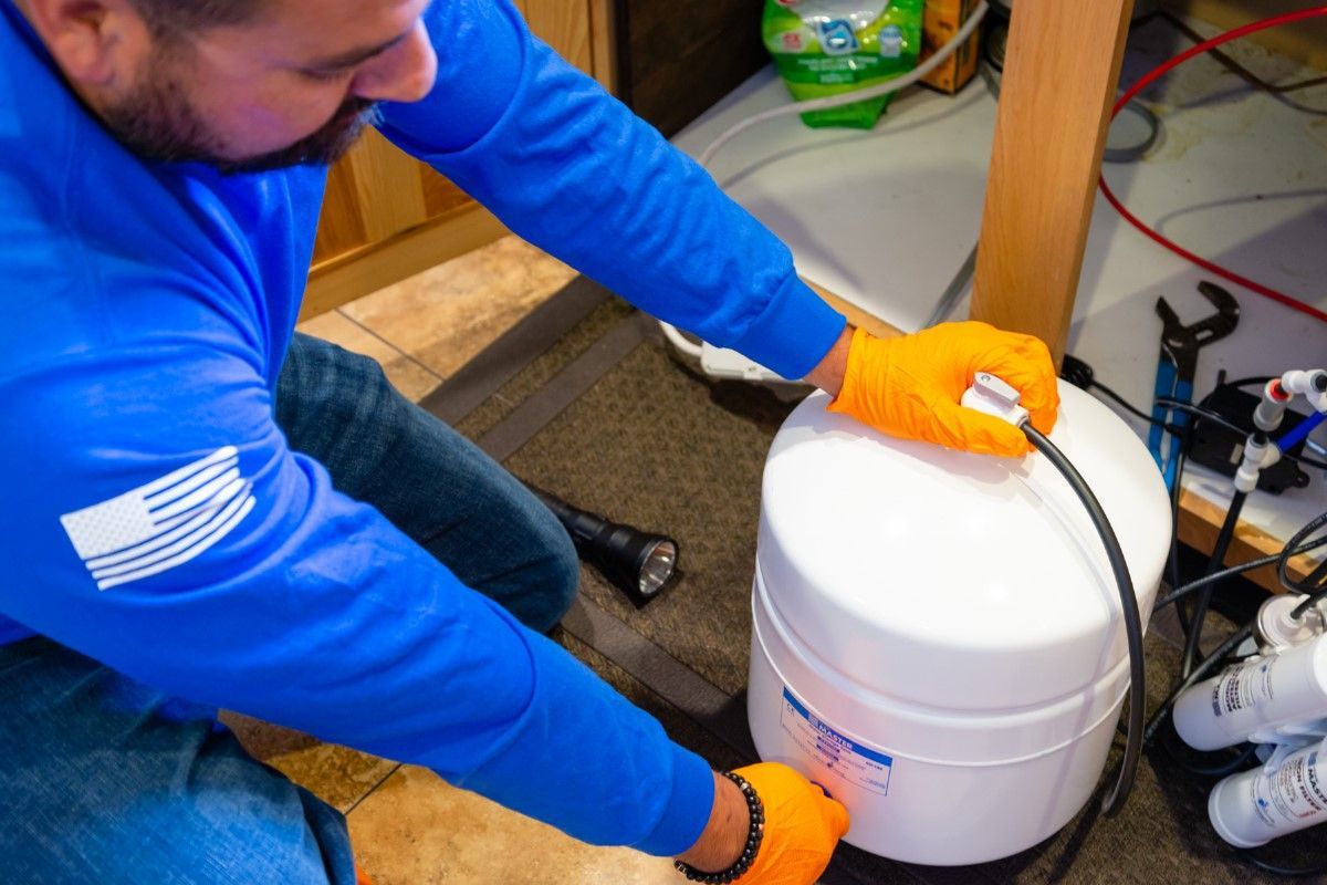 Man in blue shirt and orange gloves, kneeling, working on a white water tank.