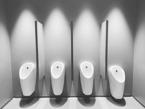 Four white urinals in a public restroom, each with a dividing wall, lit by overhead spotlights.
