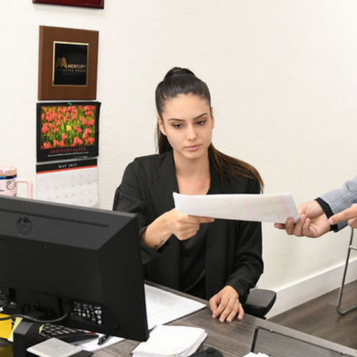 A woman is sitting at a desk in front of a computer holding a piece of paper