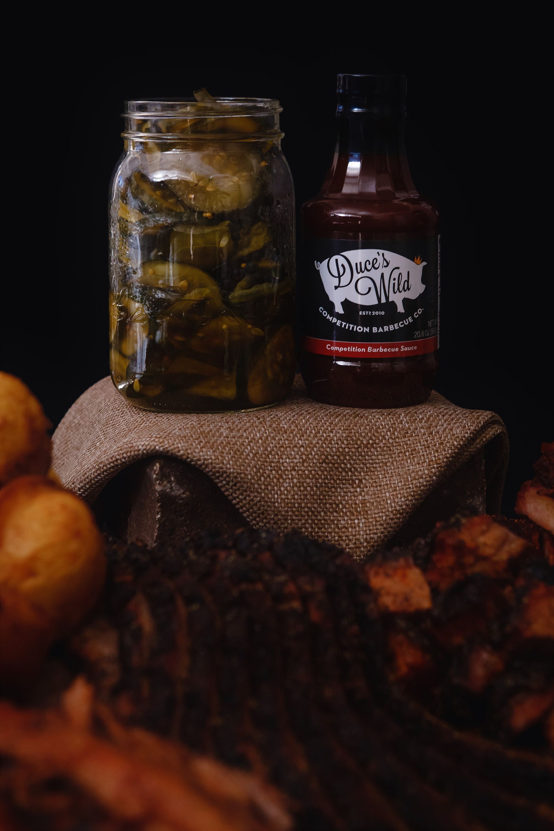 A jar of pickles and a bottle of barbecue sauce are on a table.
