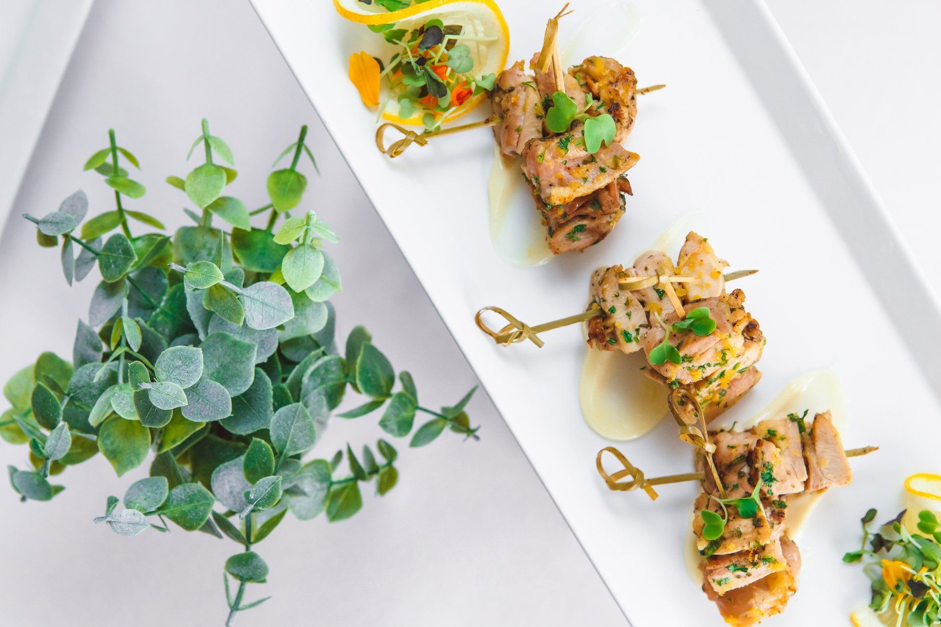 A white plate topped with skewers of meat and vegetables next to a plant.