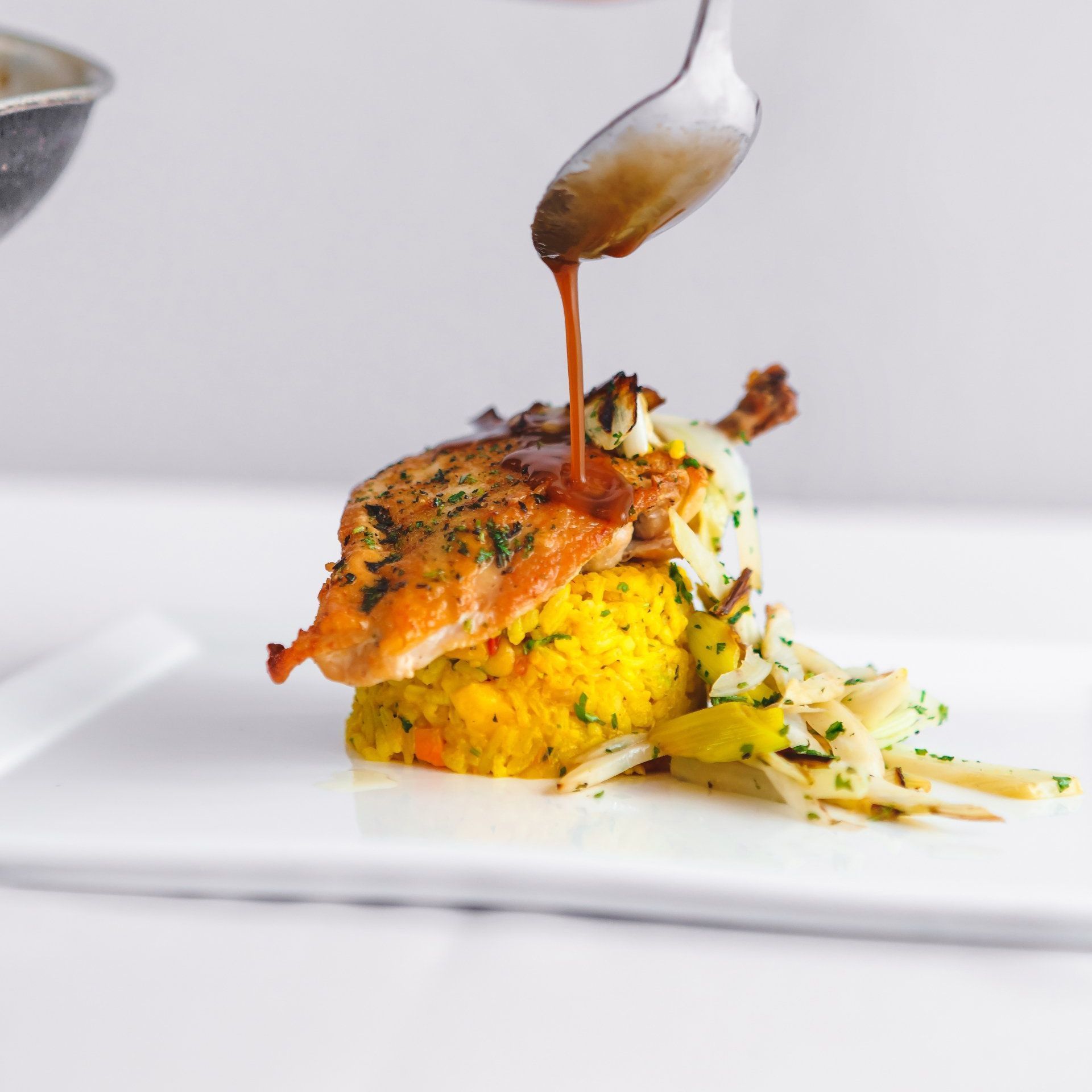 A spoon drizzles sauce over a piece of chicken on a white plate.