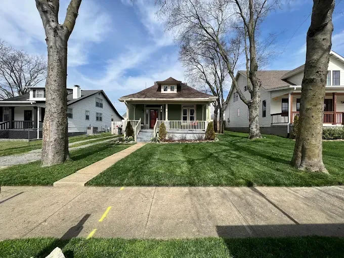 Green bungalow home with porch between two trees; sunny day.