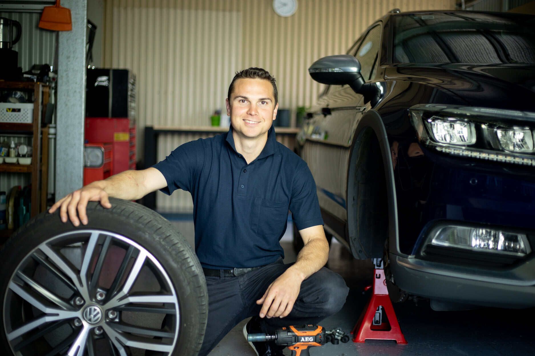 A Man is Kneeling Next to a Car Wheel in a Garage — Unified Mechanical – Mobile Mechanic In Goonellabah, NSW