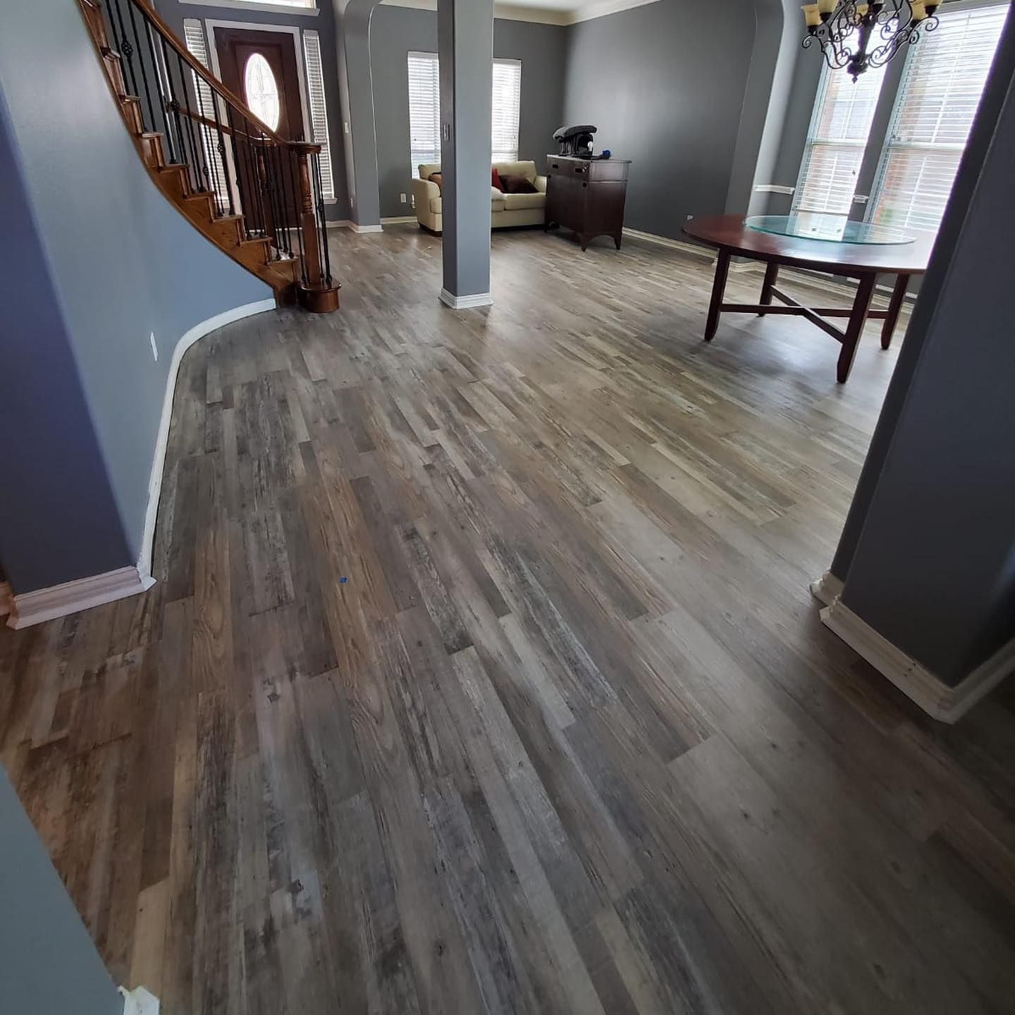 Wooden floor in a home's entryway, leading to a staircase and dining area. Gray walls, dark furniture, and a chandelier.