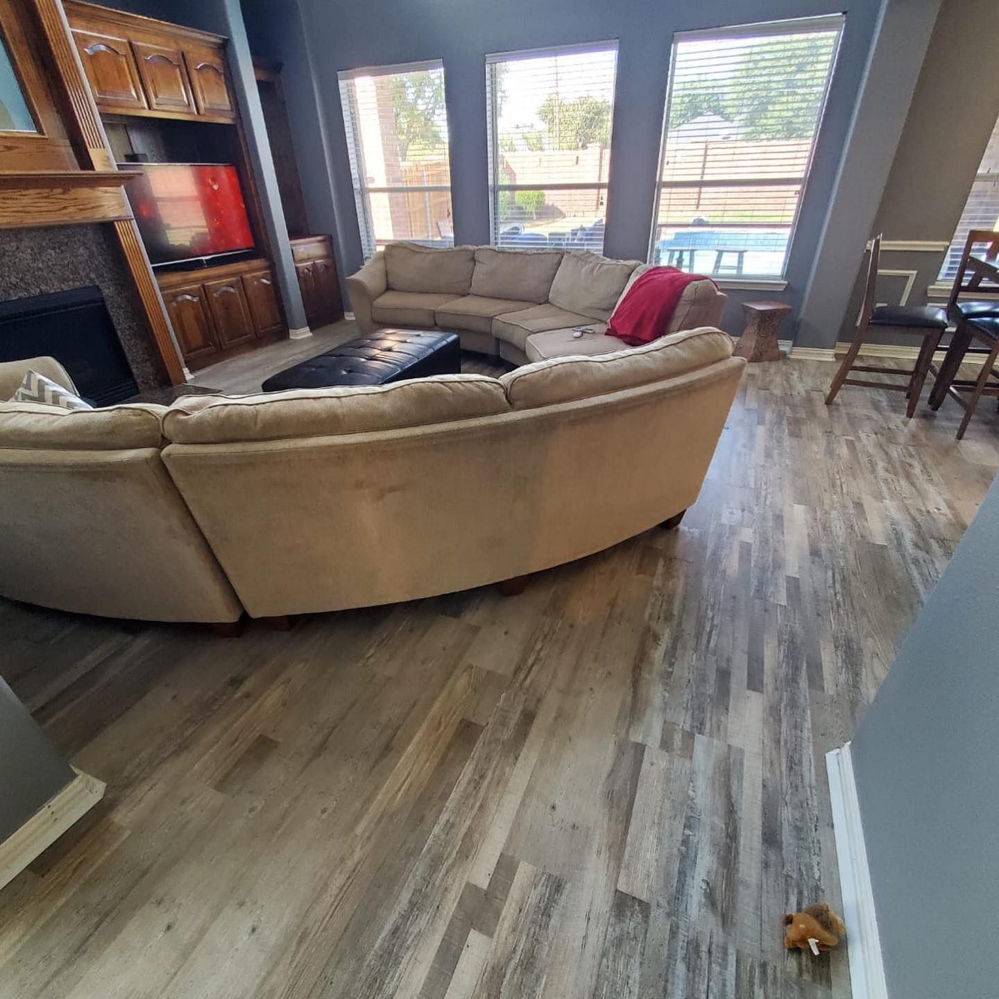 Living room with curved beige sofa, fireplace, TV, and wood-look flooring. Windows overlook a backyard.