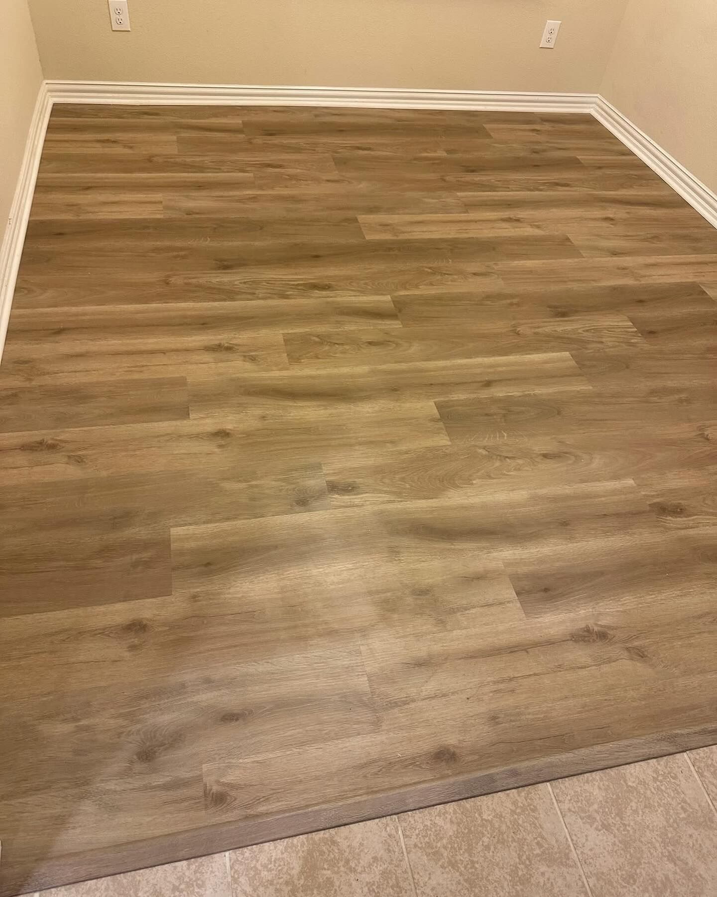 Brown wood-look flooring fills a room, with white trim along the walls, a neutral carpet border is visible.