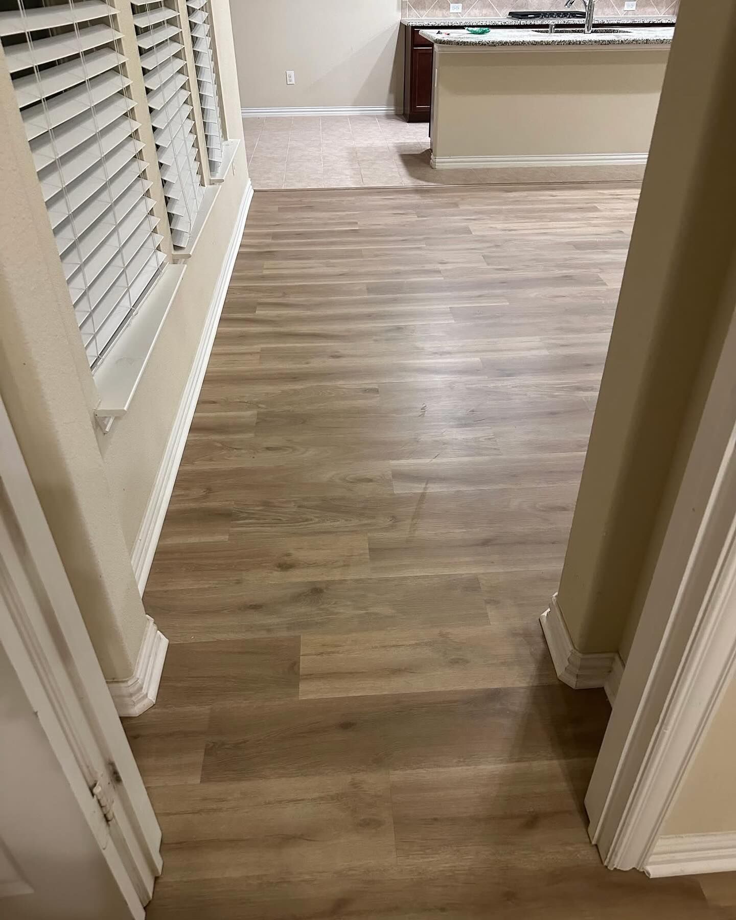 Wooden floor leading to a kitchen, beige walls, and a window with blinds.