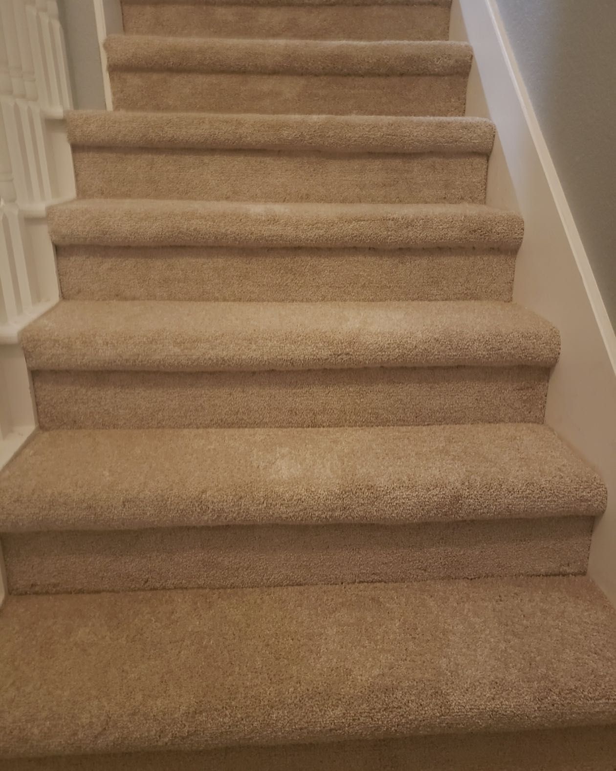 Carpeted staircase, neutral tan color, ascends upwards against a white wall and railing.