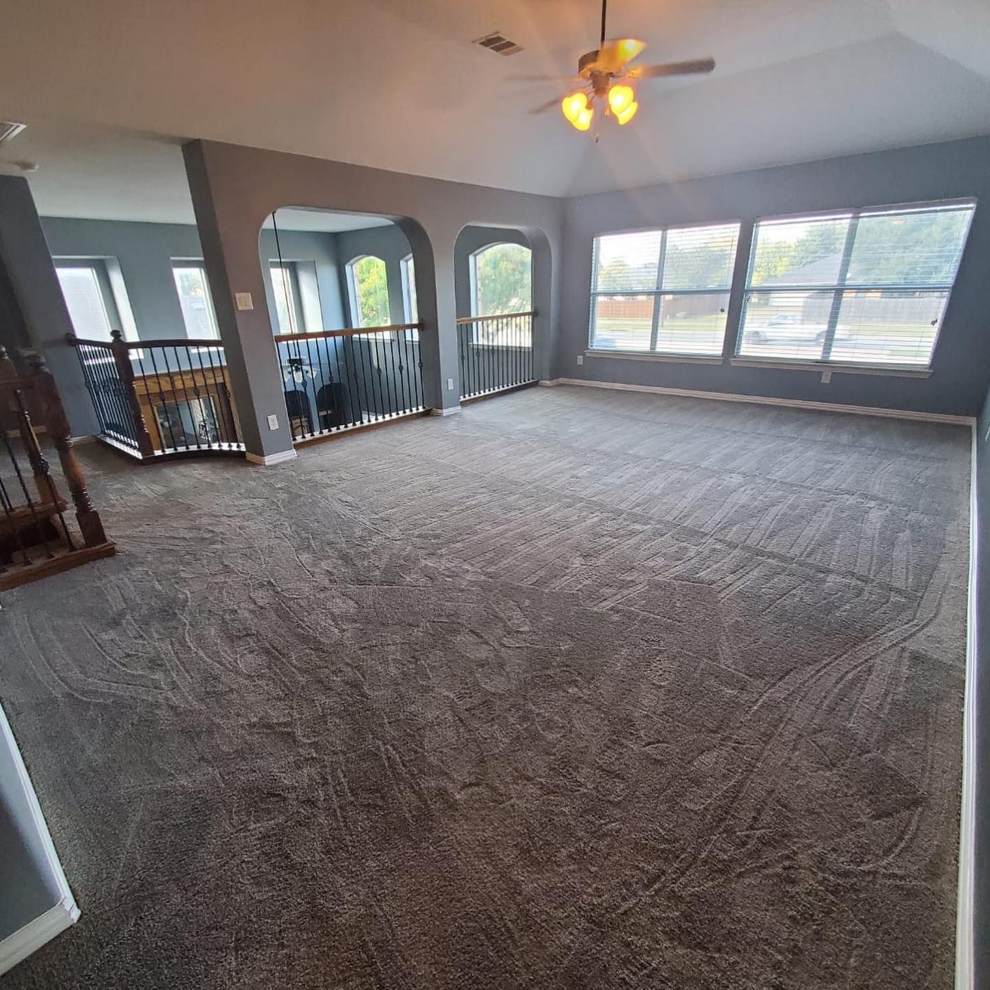Spacious living room with gray carpet, walls, and large windows. A staircase and second-floor railing are visible.