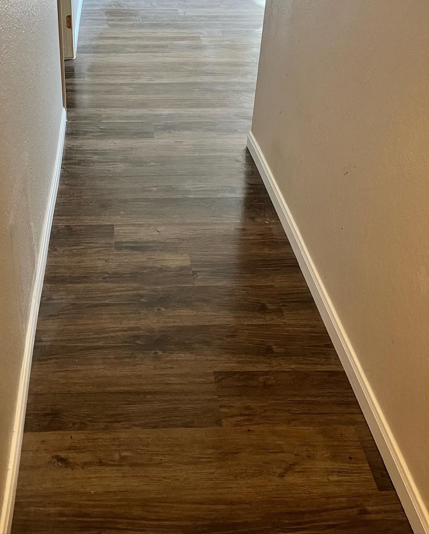 Narrow hallway with dark wood-look flooring and beige walls. White trim lines the floor edges.