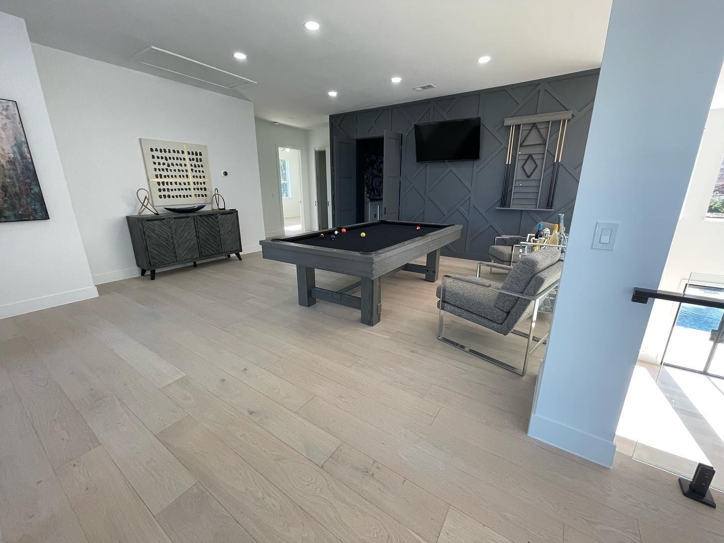 A modern game room with a pool table, gray wall, and light wood floors.