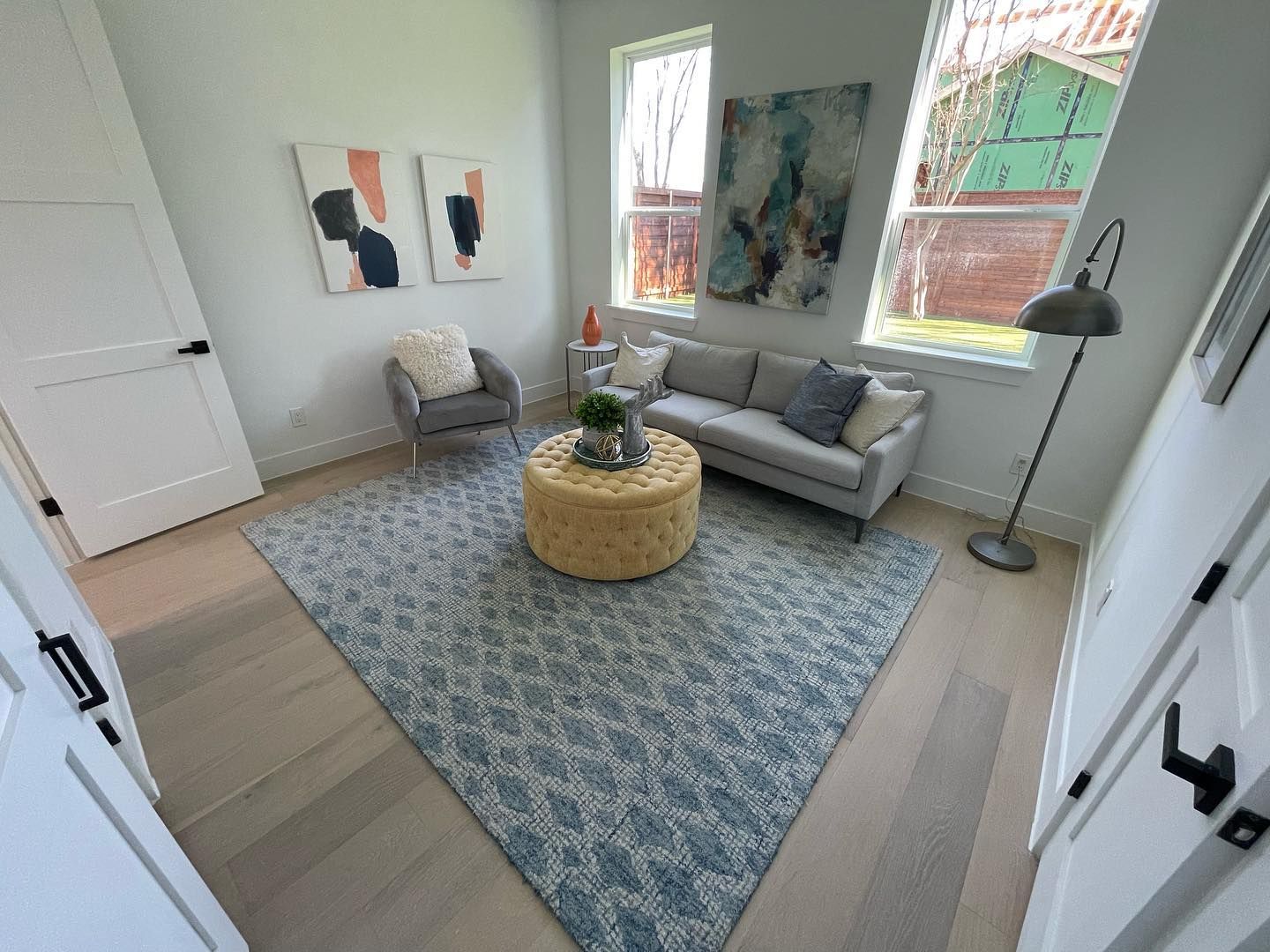 Living room with a blue rug, gray sofa, two paintings, and a round ottoman.