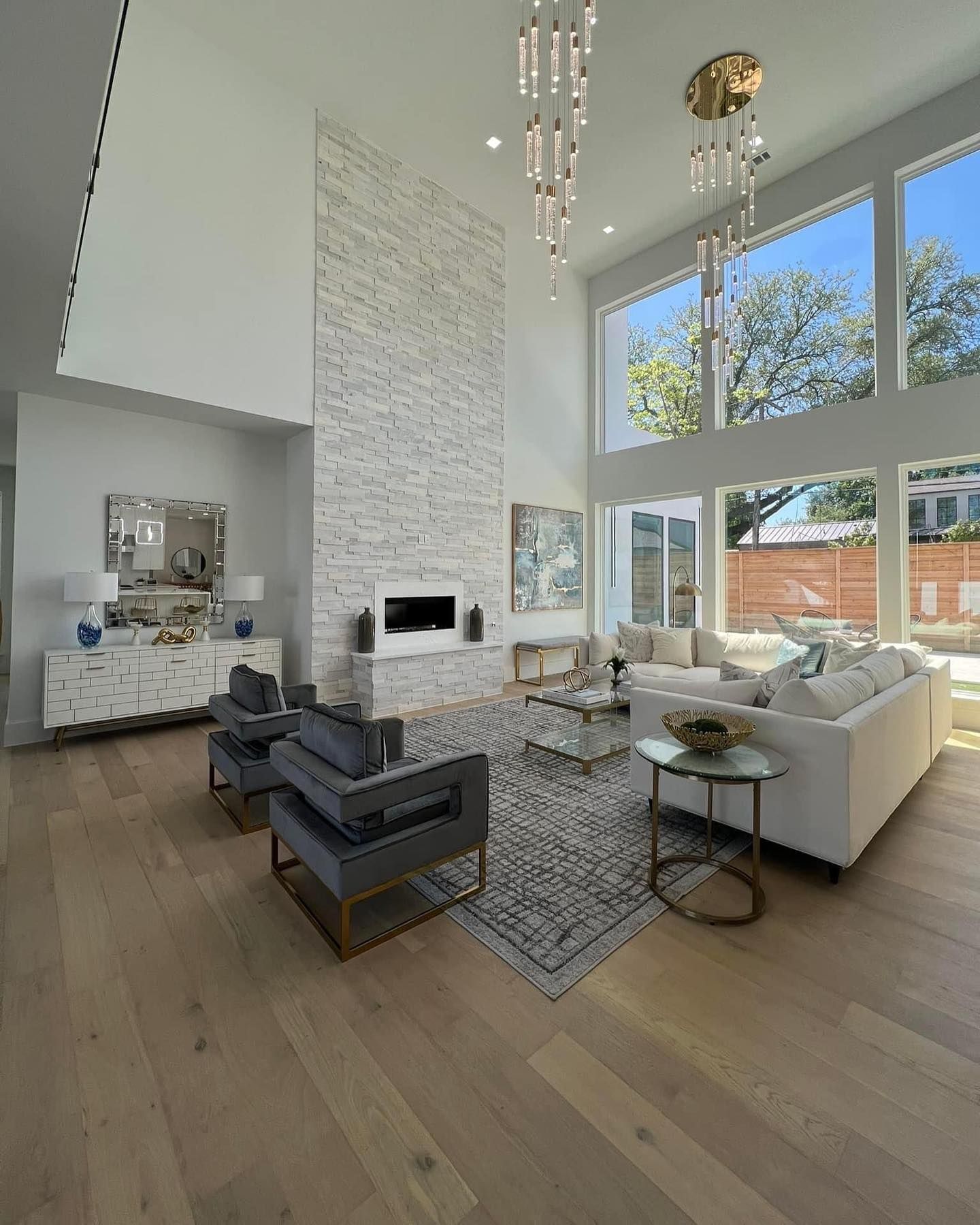 Modern living room with high ceilings, light wood floors, white stone fireplace, and large windows.
