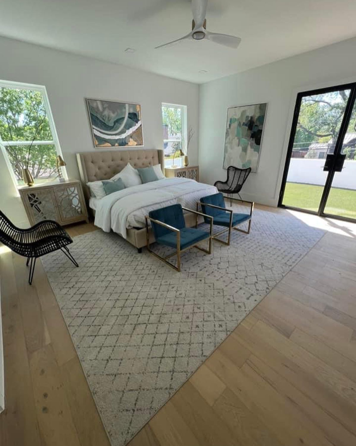 Spacious, modern bedroom with light wood floors, large rug, king bed, teal chairs, and large windows overlooking yard.