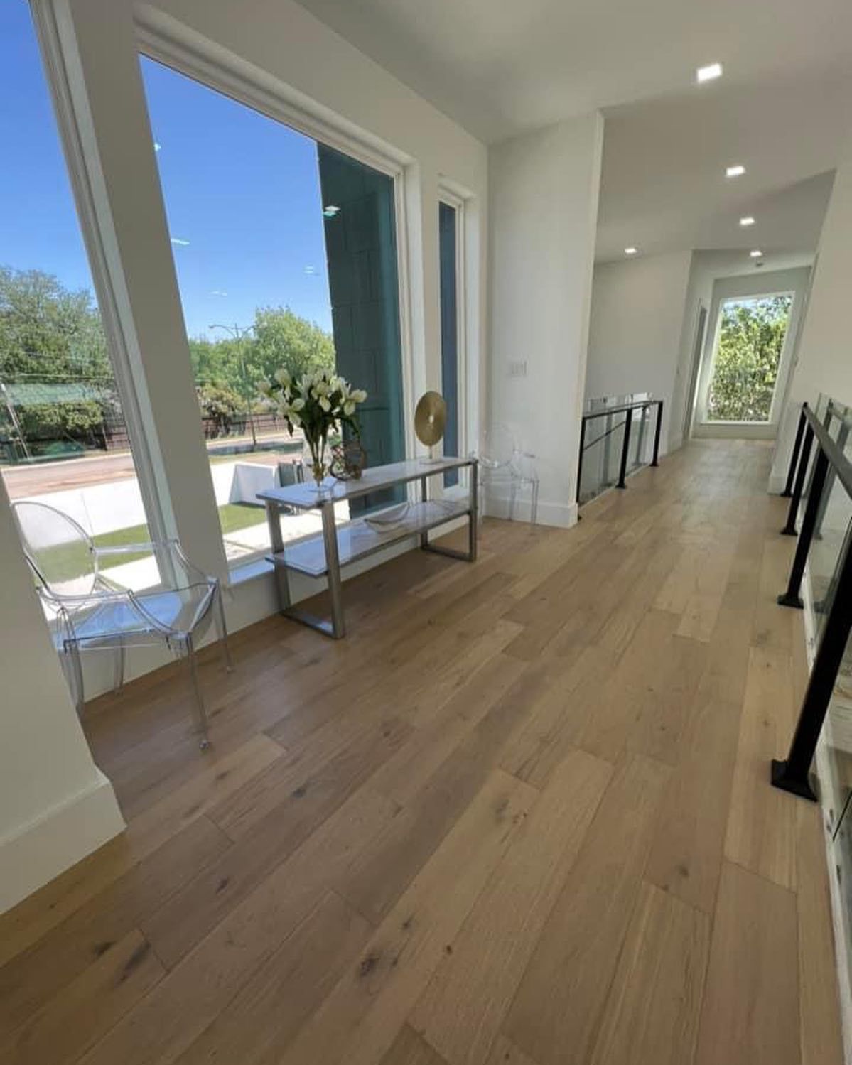 Hallway with light wood floors, large window, white walls, and a table with flowers.