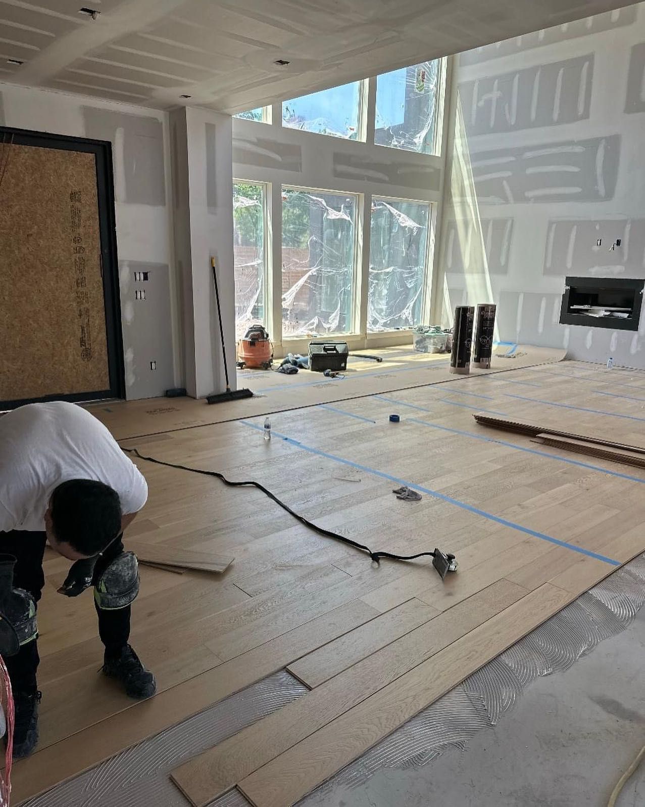 Person installing hardwood flooring in a room with large windows. Construction site.