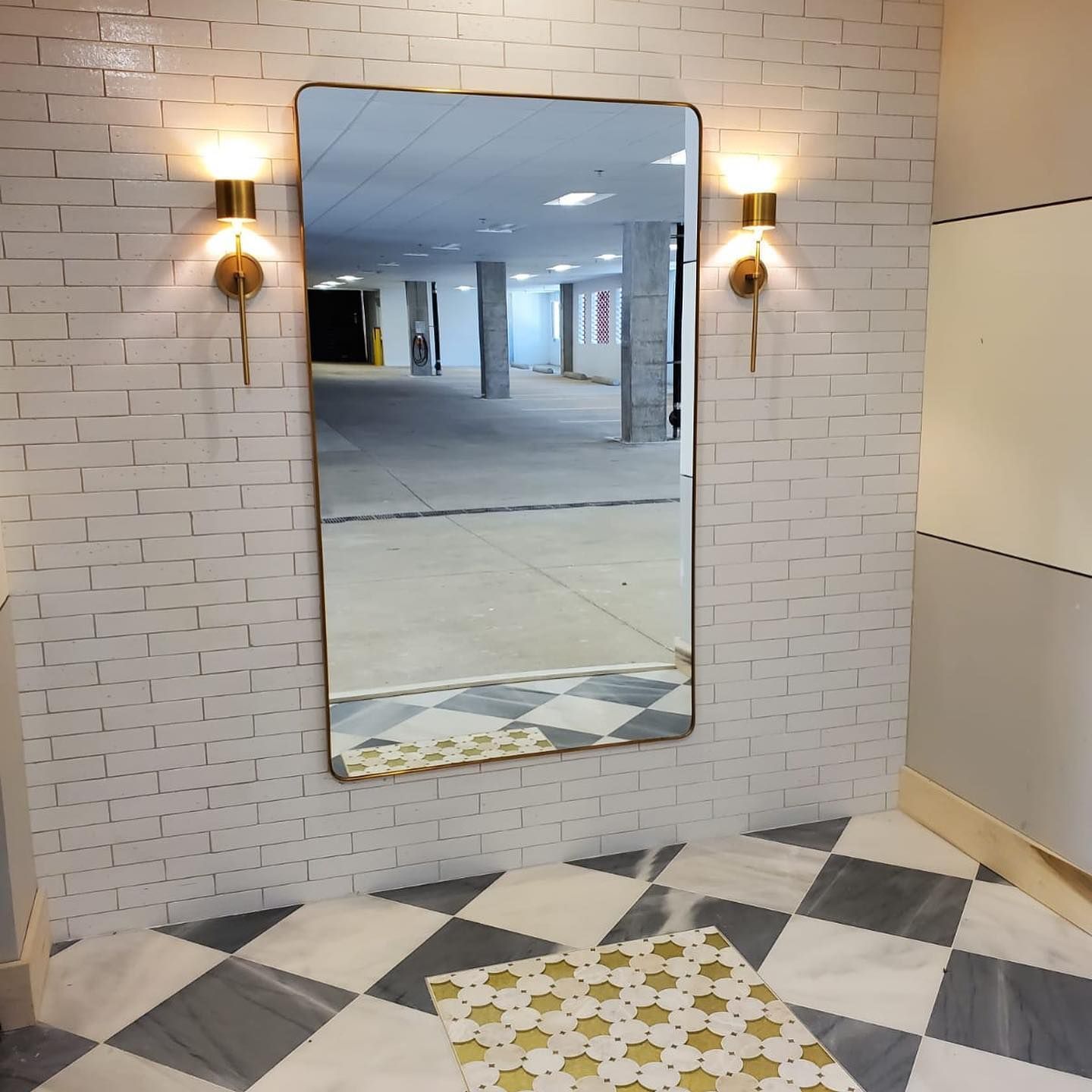 Mirror reflecting an empty parking garage, flanked by sconces on a white brick wall, over a checkered floor.