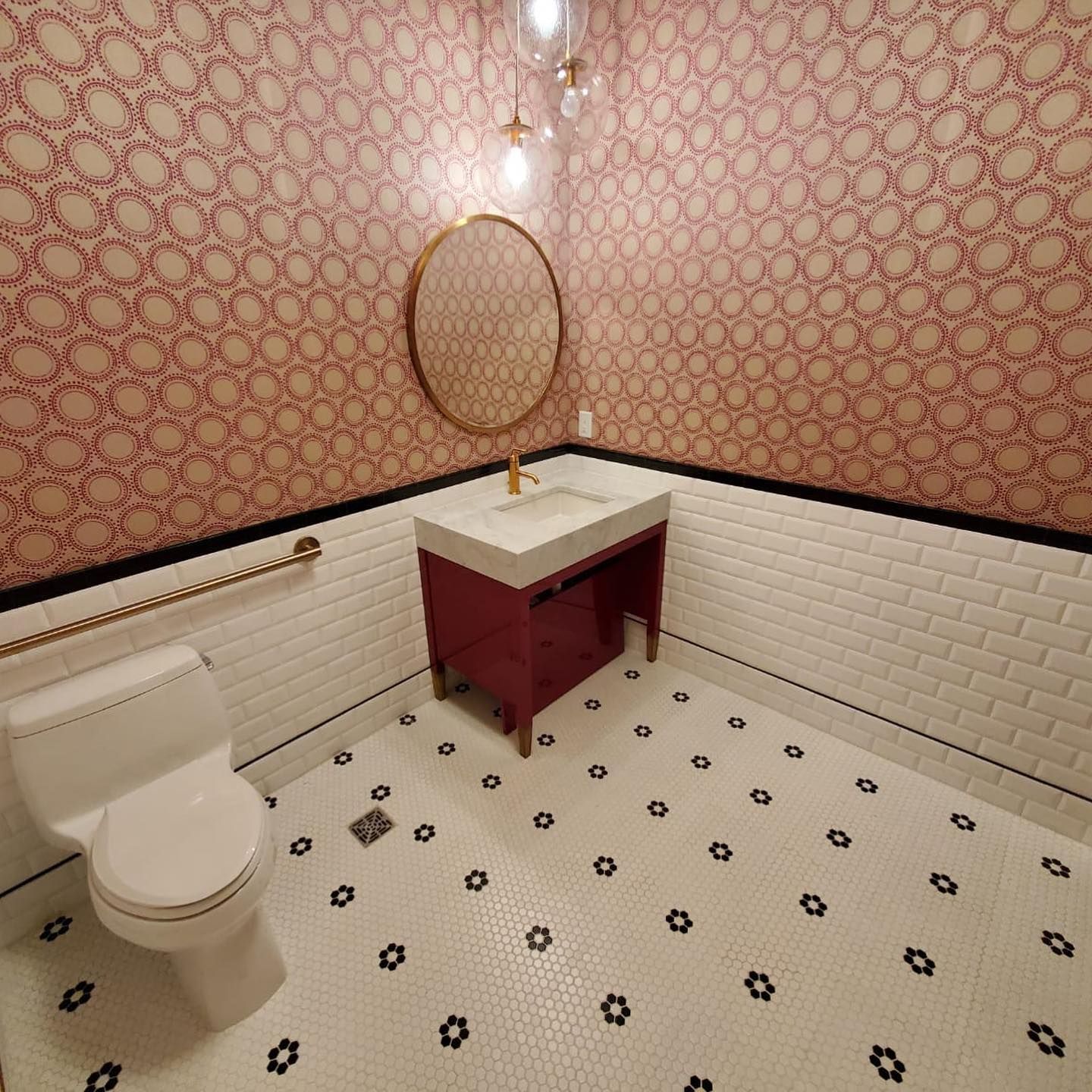 A small bathroom with pink wallpaper, white tile, and a red vanity.