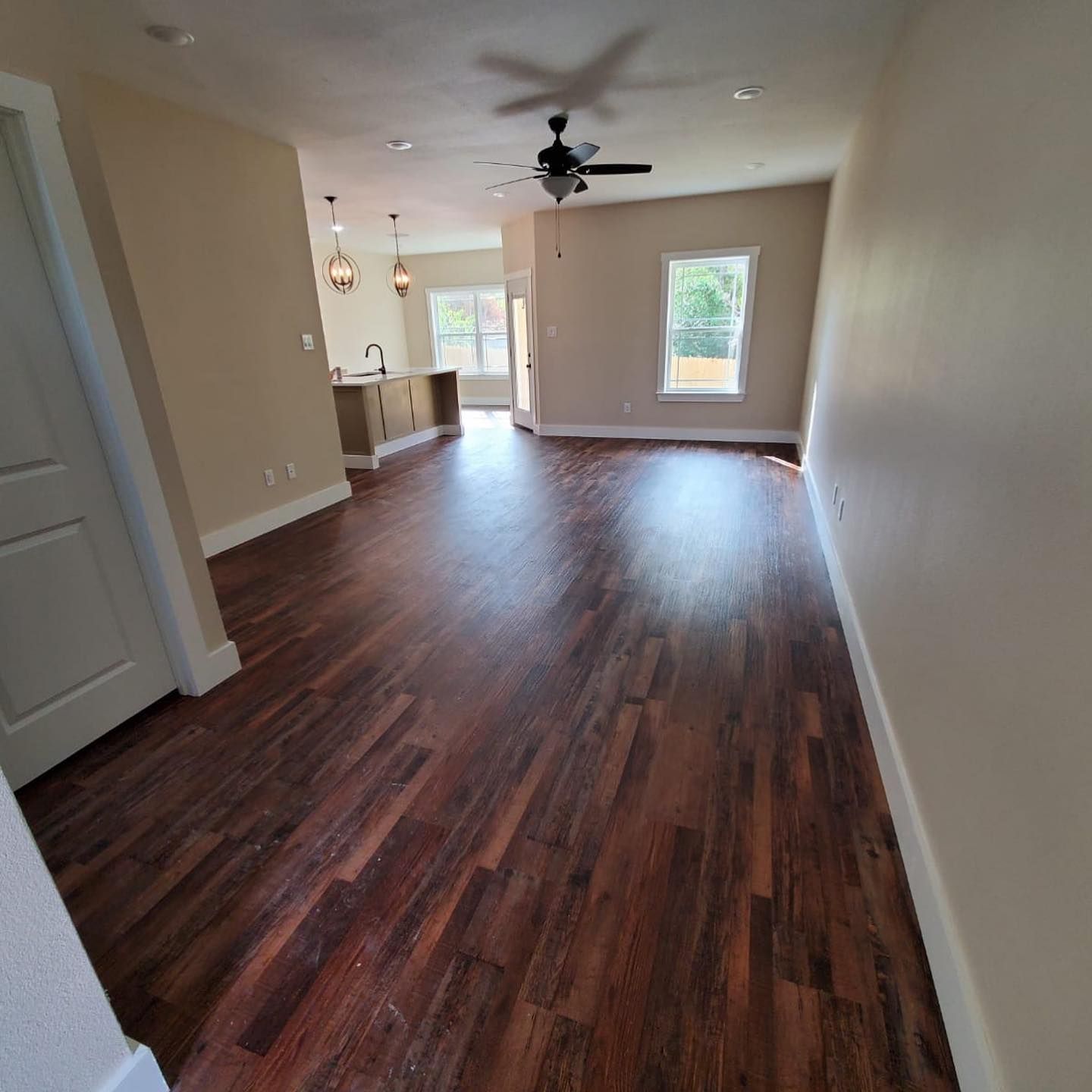 Interior view of a living space with wood flooring, tan walls, and a kitchen in the background.