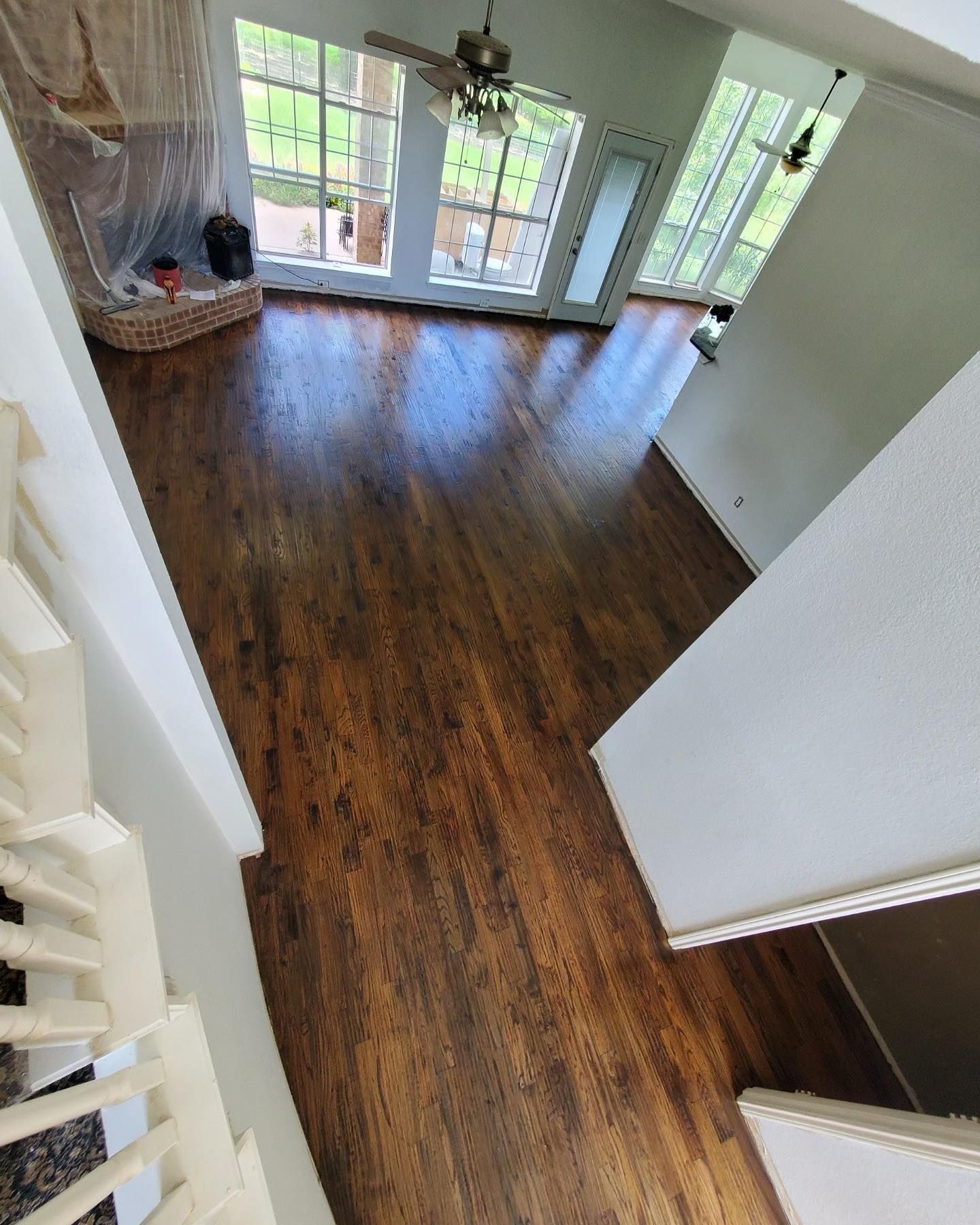Dark-stained wood floor in a room with large windows, seen from a staircase. Bright natural light.