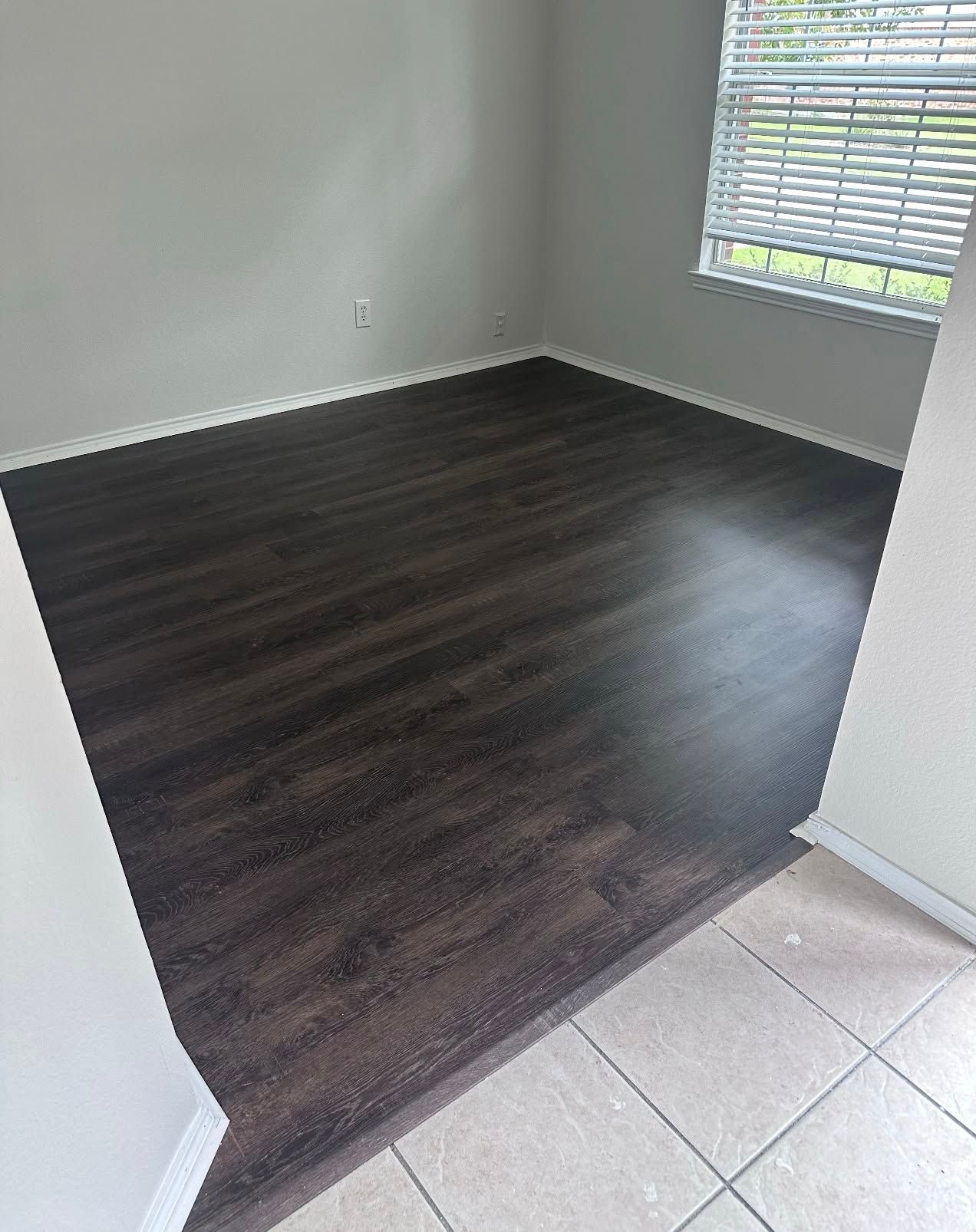 Empty room with dark wood-look flooring and a window with blinds. Beige tile floor in foreground.