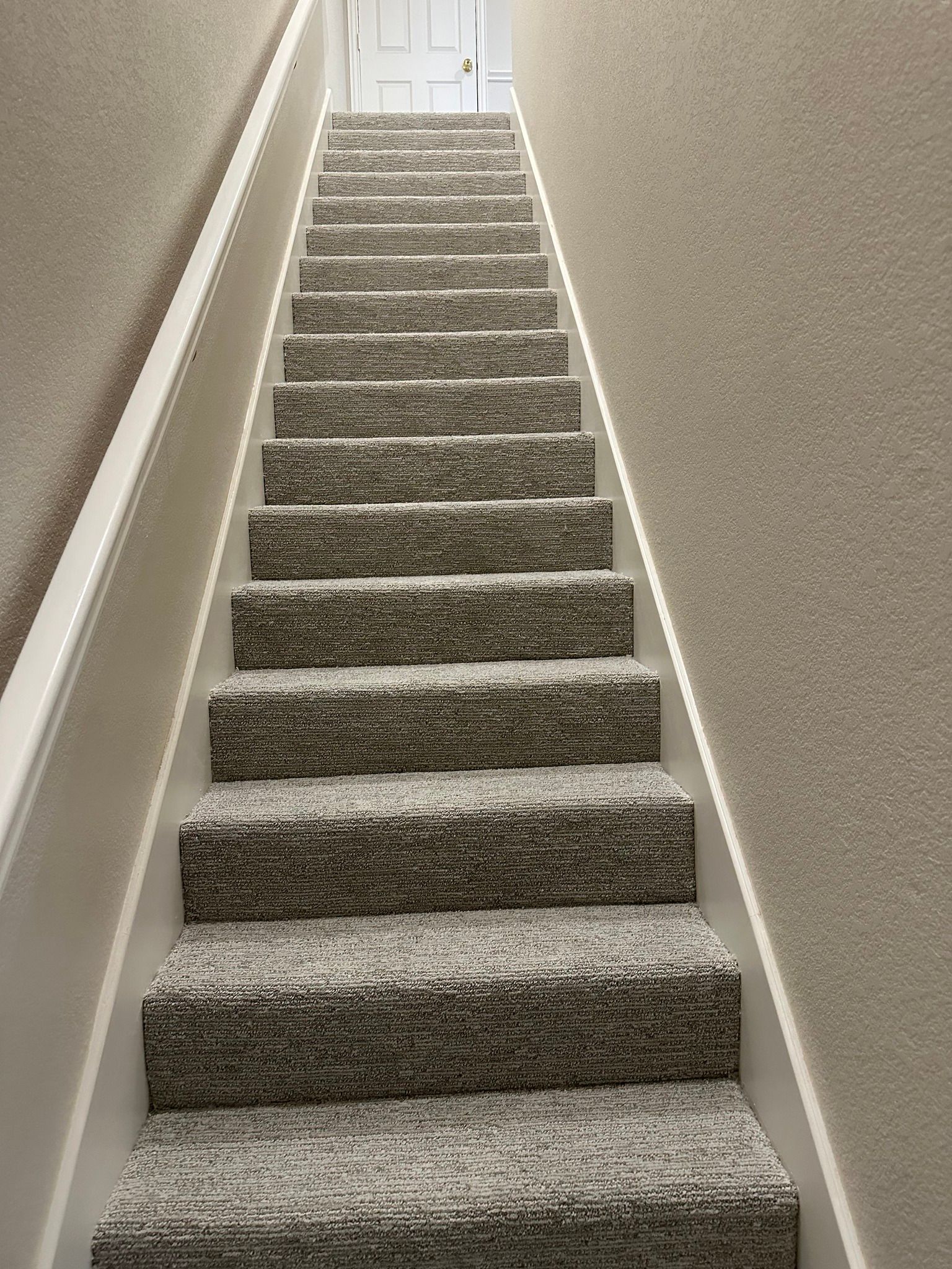 Staircase with carpeted steps and white banister. Beige walls frame the stairs, leading up to a white door.