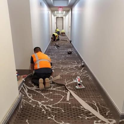 Workers installing carpet in a hotel hallway; one kneels, others work in the background.