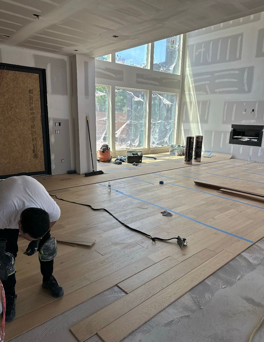 Person installing wood flooring in a room with large windows, unfinished walls, and a fireplace.
