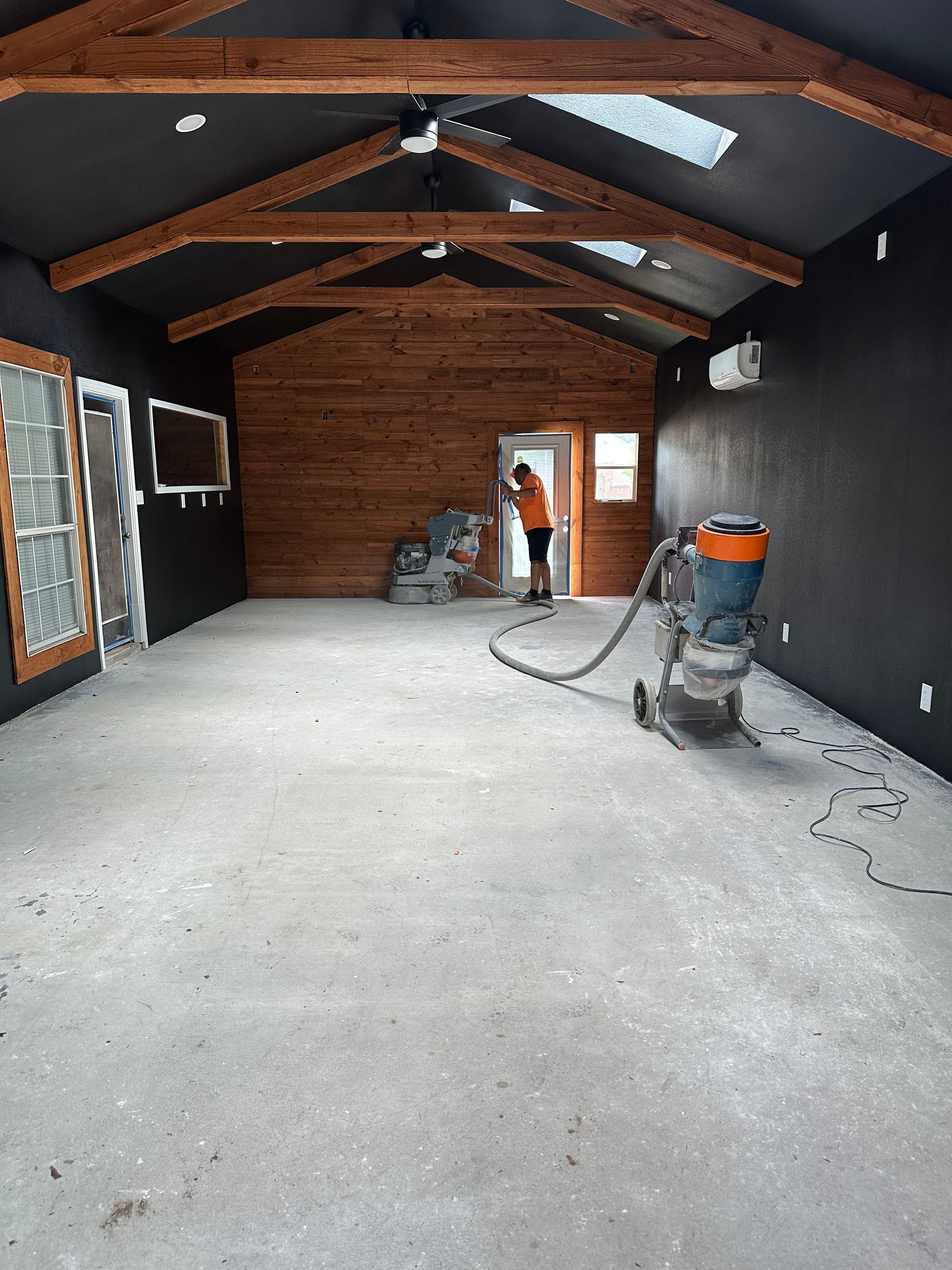 Interior view of a room with a person using a floor sander. Concrete floor, wood beams, and dark walls.