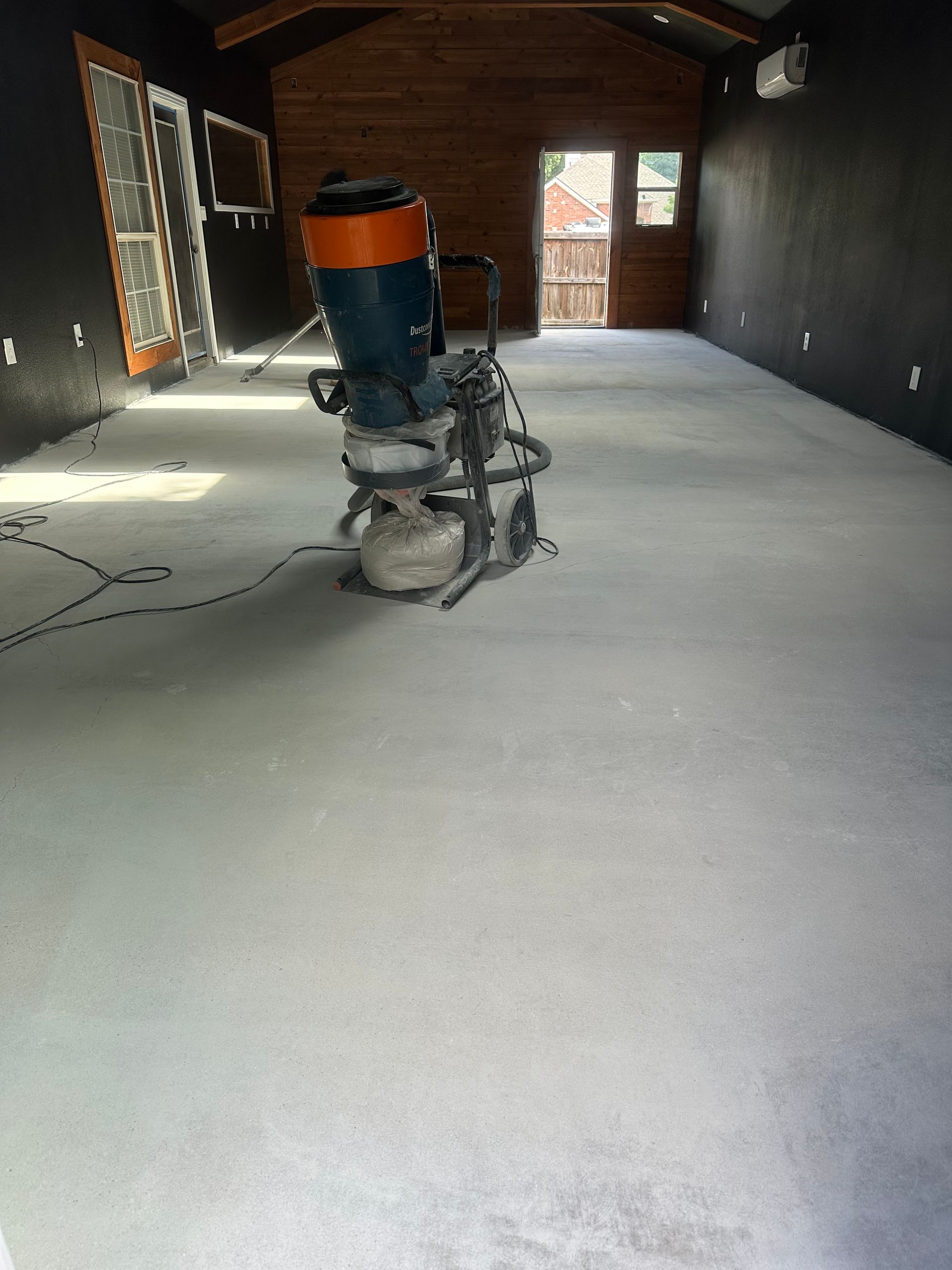 Concrete floor being ground by an industrial floor grinder in a room with dark walls and a wooden door.