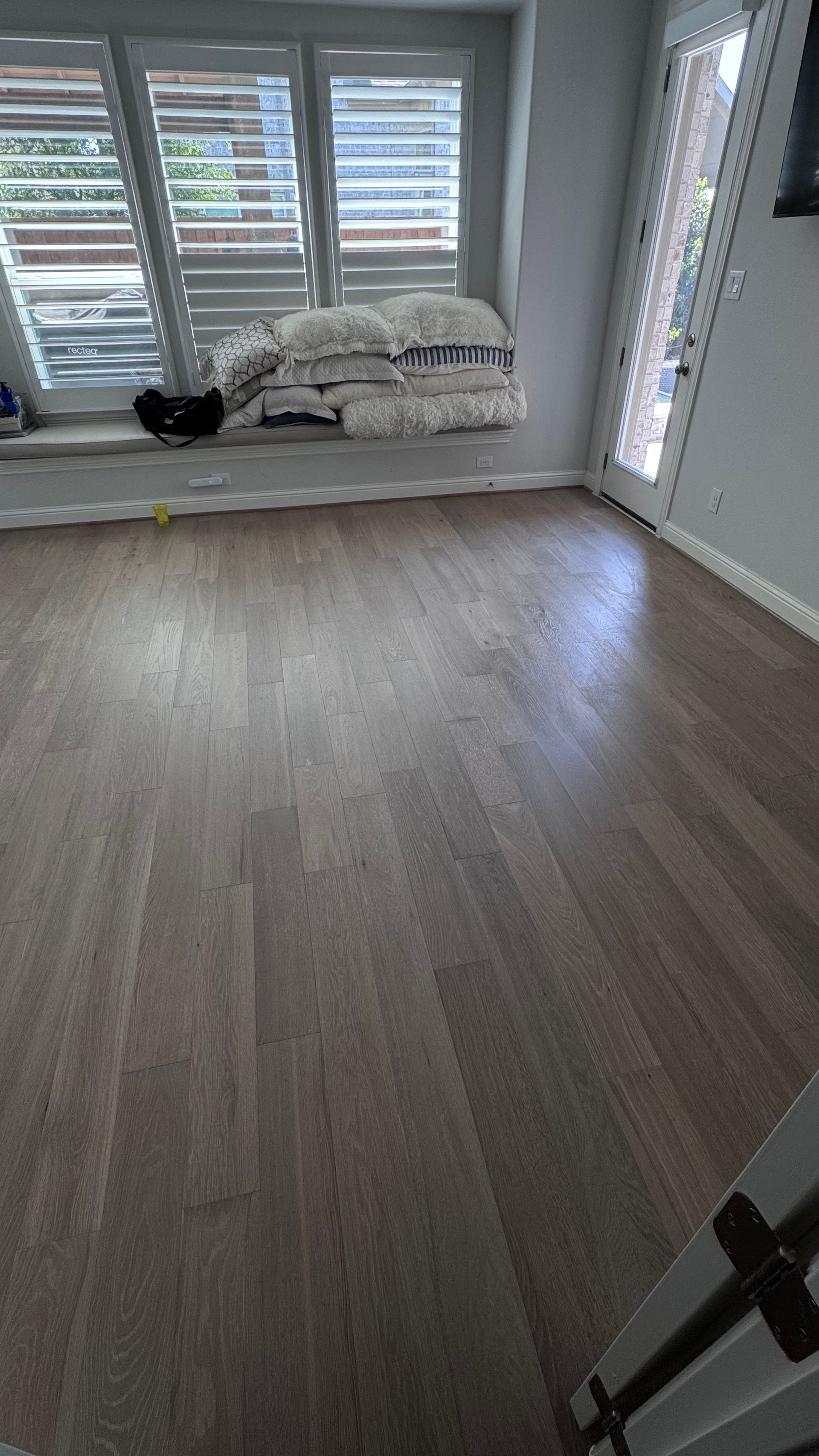 Newly installed gray wood flooring in a room with a window, door, and shuttered window in the background.