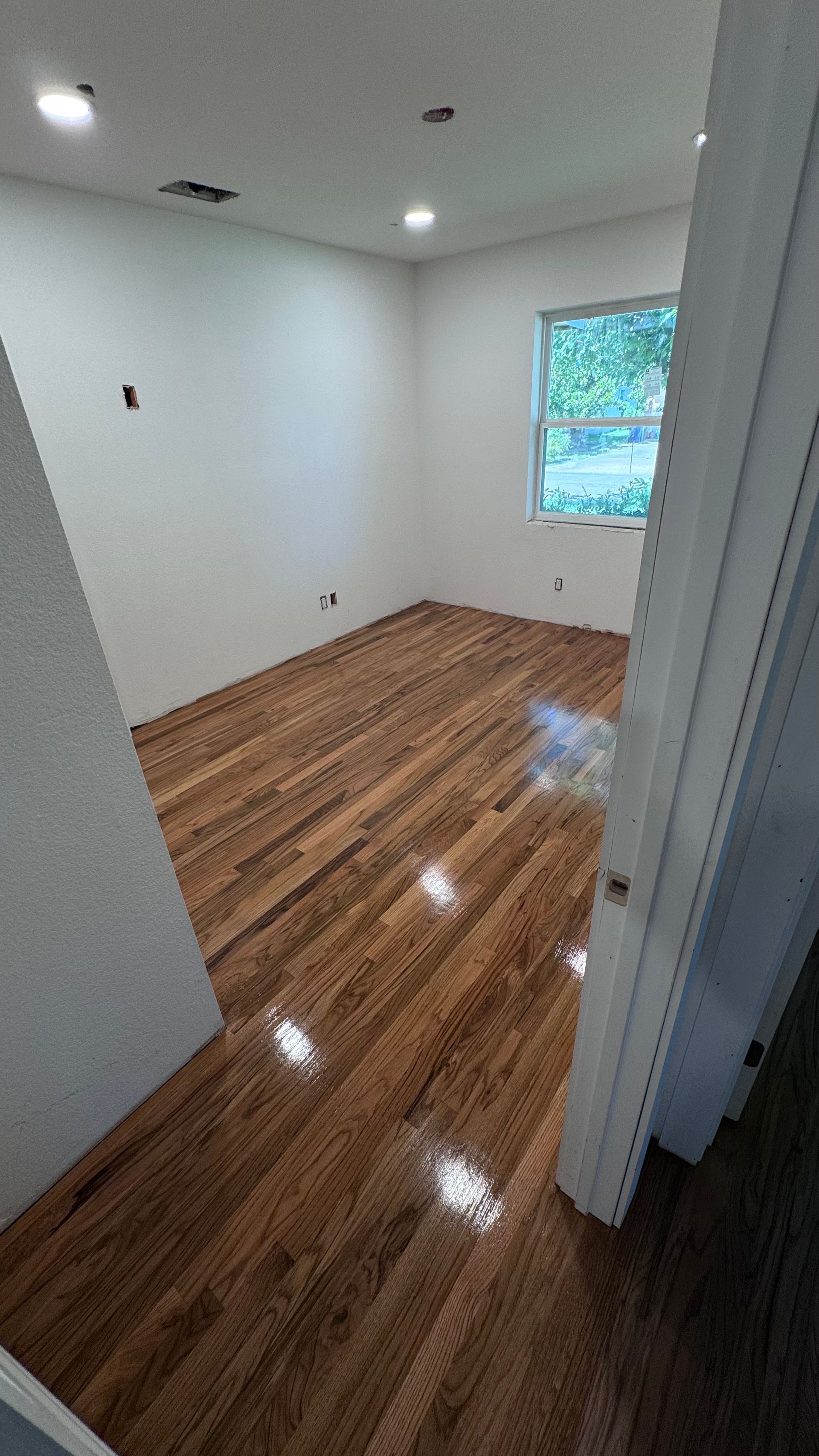 Empty room with hardwood floors, white walls, and a window with sunlight.