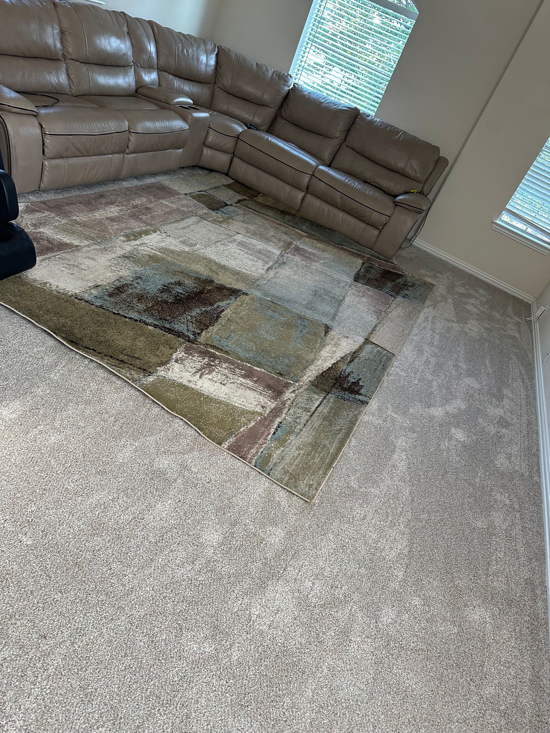 Tan sectional sofa on a patterned rug over light gray carpet. Two windows with blinds in the background.