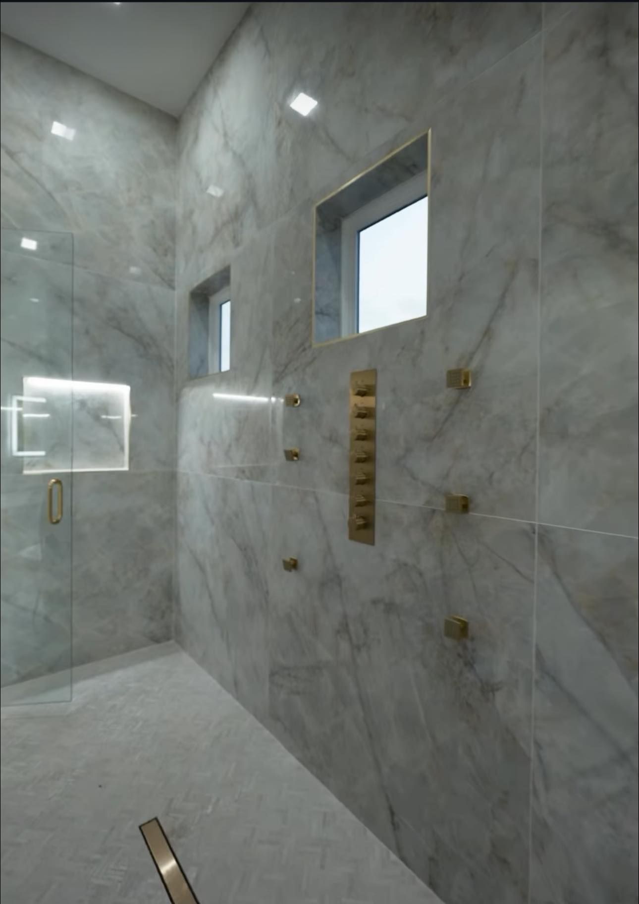 Luxurious marble-tiled shower with gold fixtures, glass enclosure, and natural light from windows.