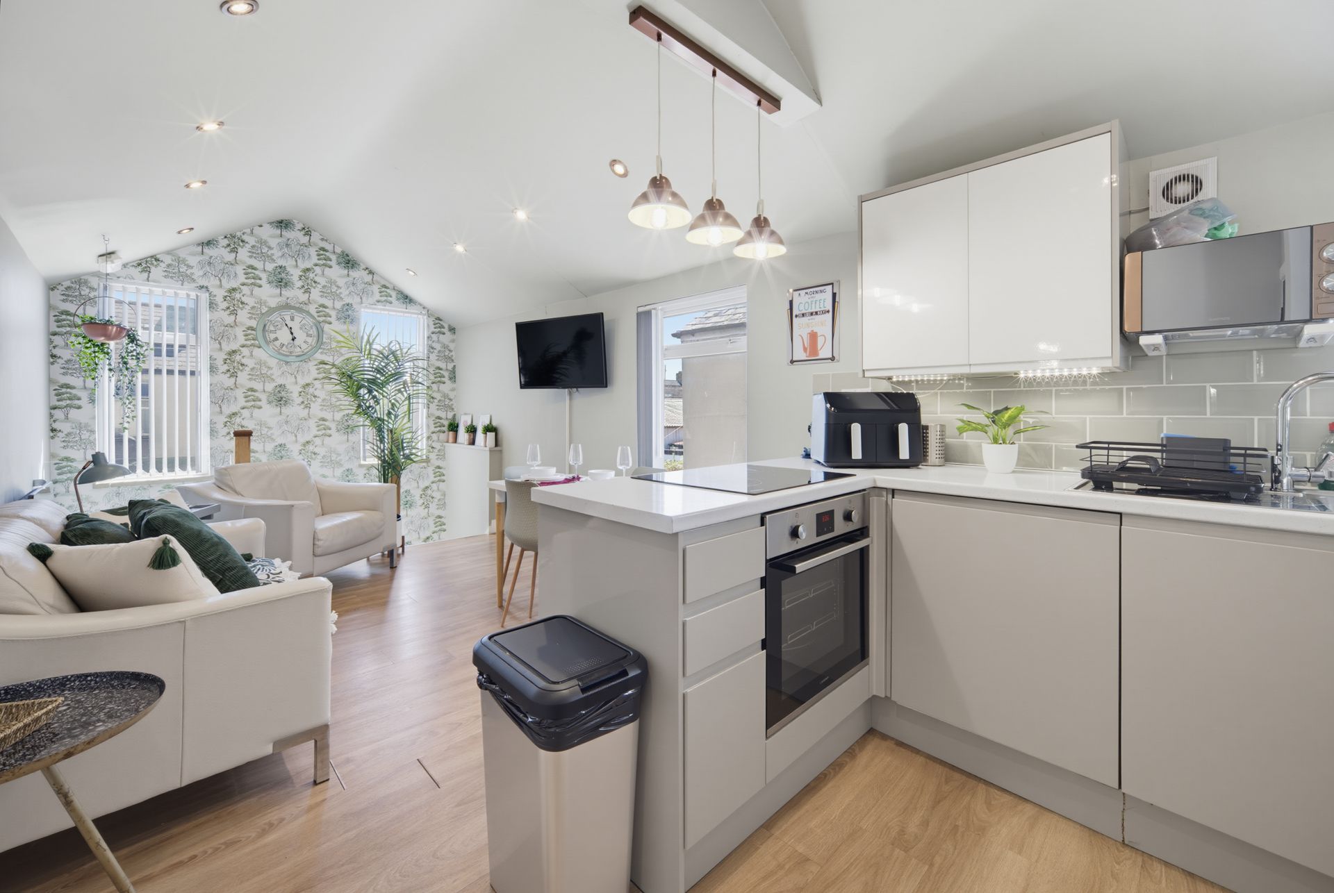 Bright open-plan kitchen and living room with white cabinets, gray sofa, and large windowed wall.