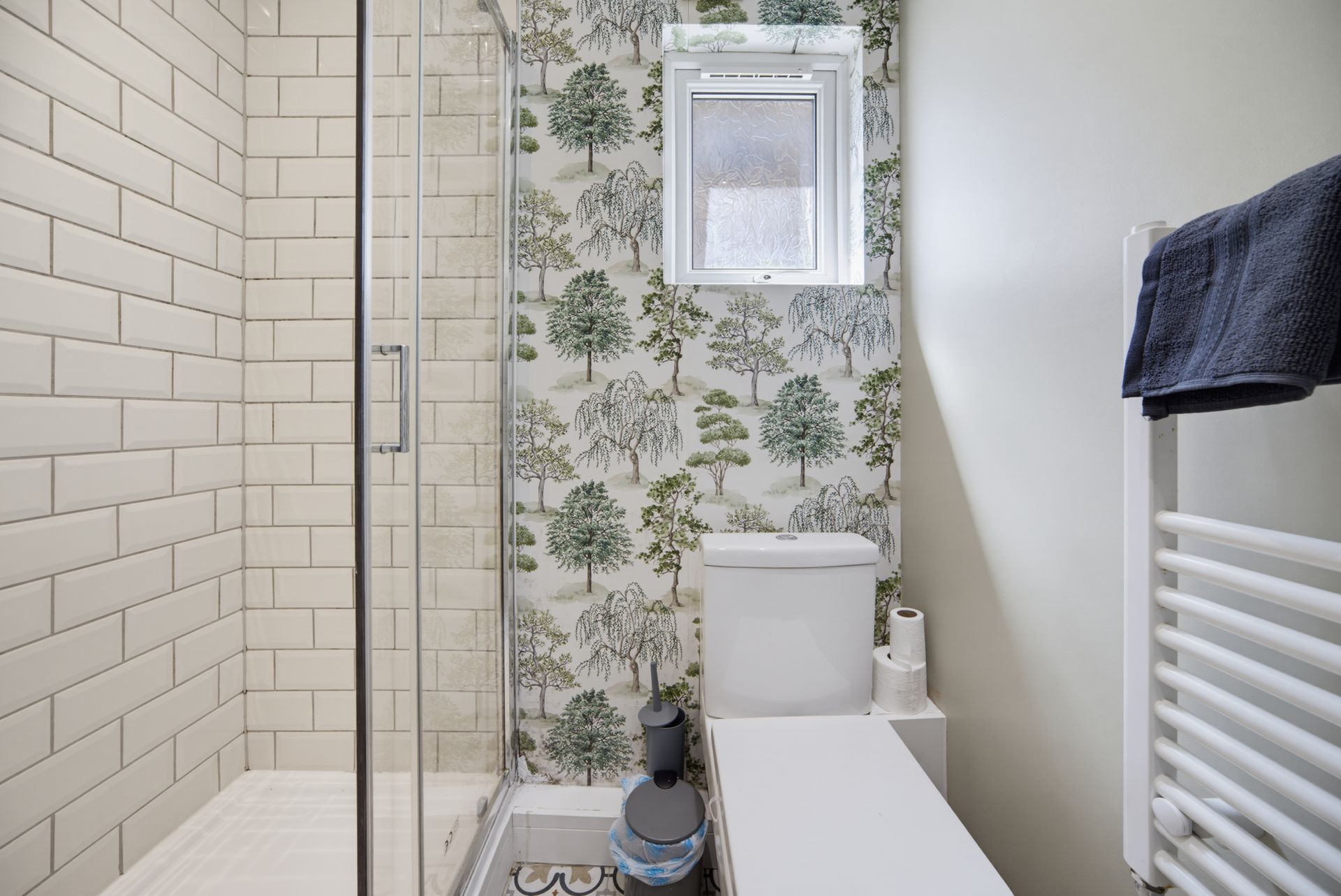 Small bathroom with glass shower, toilet, floral wallpaper, white radiator, and window.