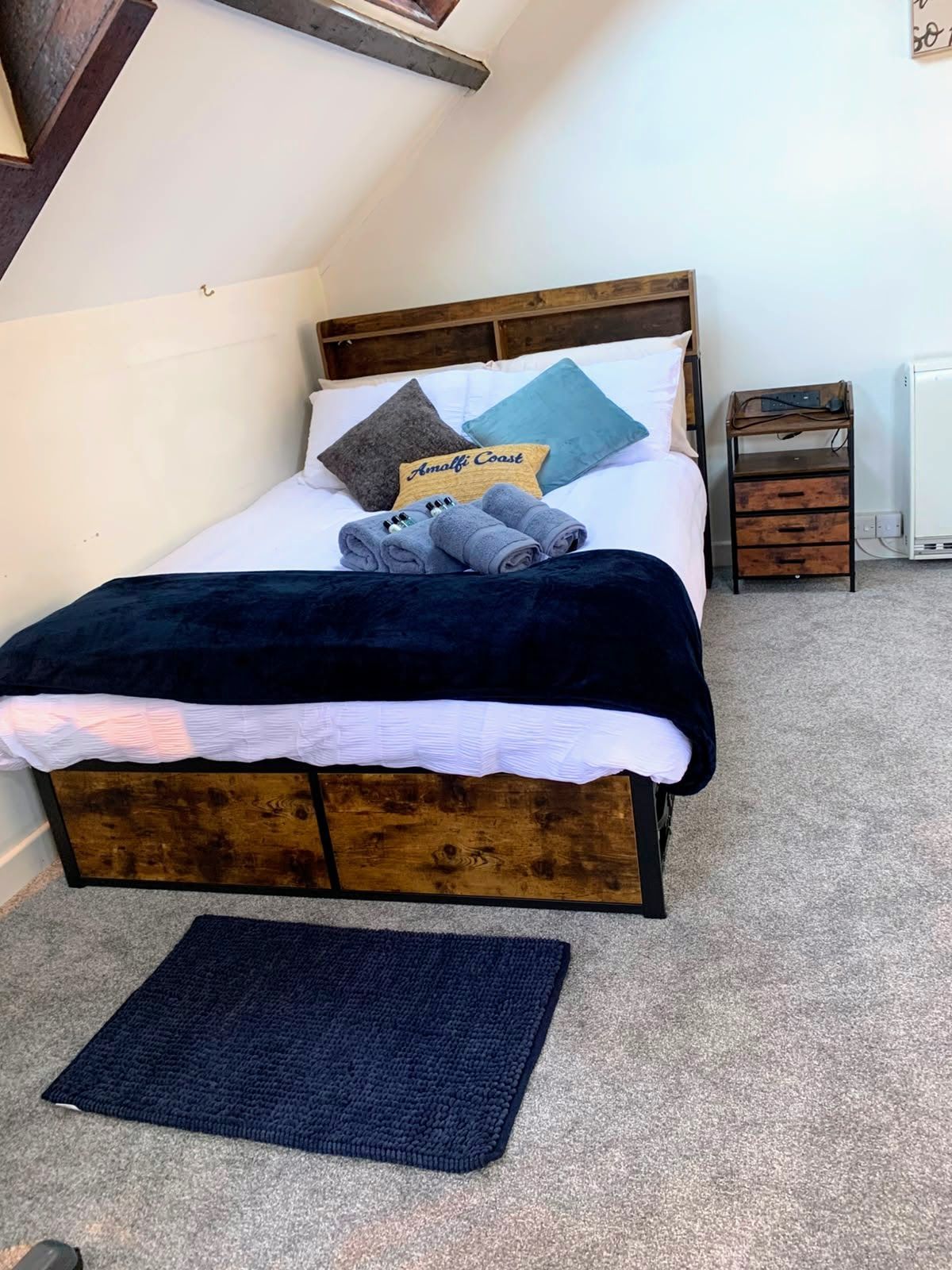 Small attic bedroom with a wooden bed, blue bedding, and a dark rug on carpeted floor.