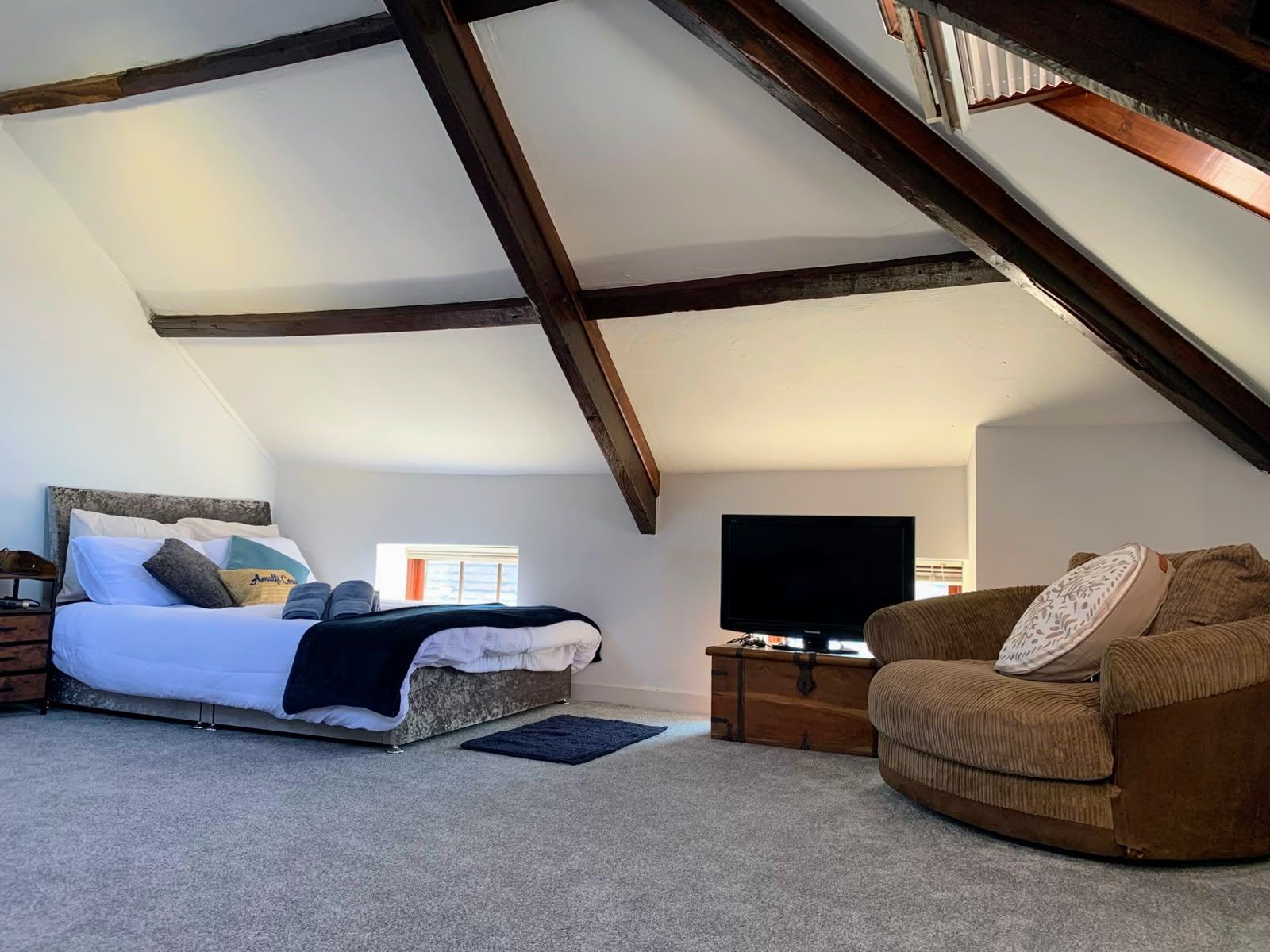 Cozy attic bedroom with exposed beams, a bed, TV, and wicker chair on gray carpet