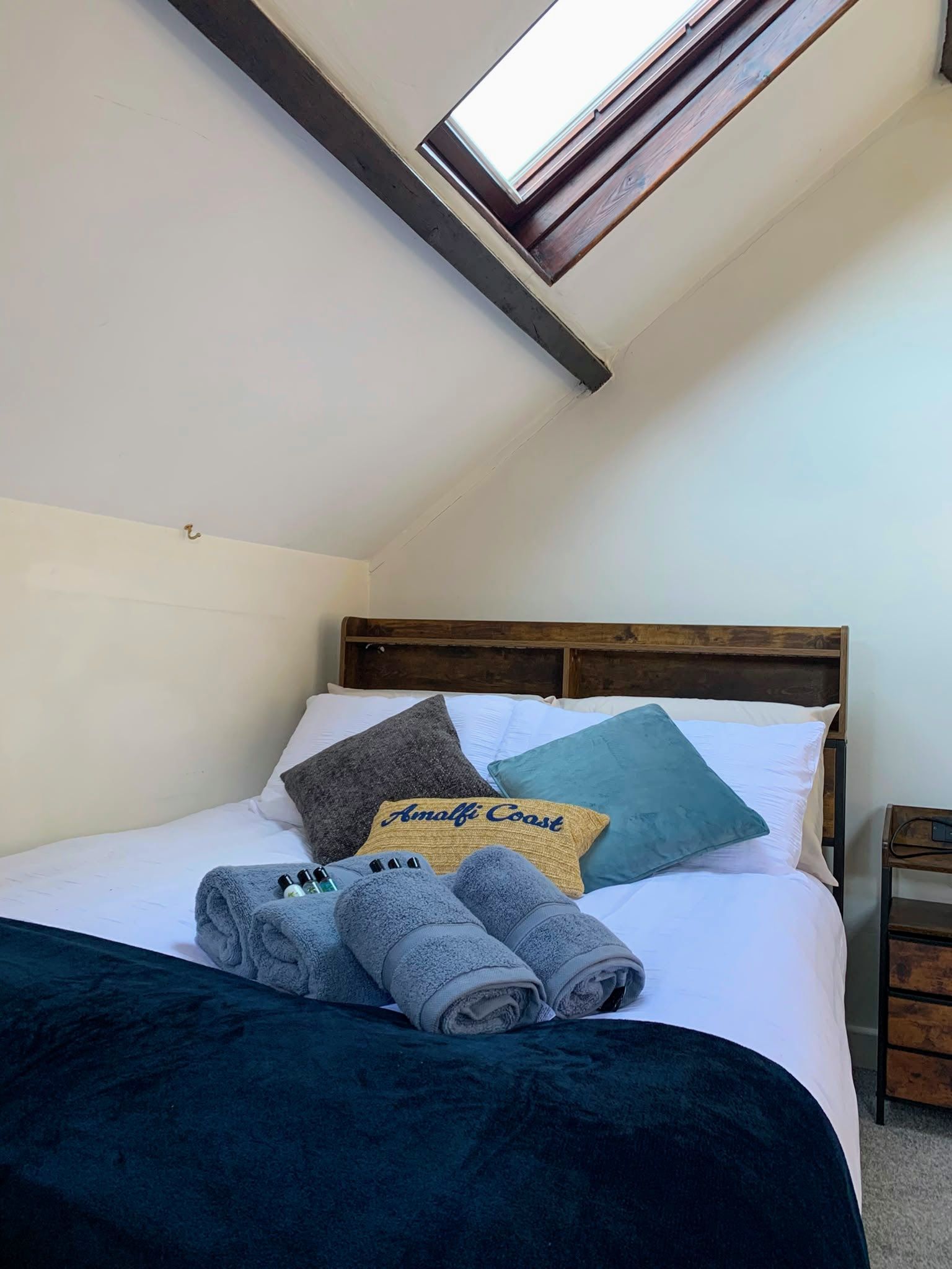Cozy bed under a skylight, with white bedding, teal pillows, and folded towels in a small attic bedroom